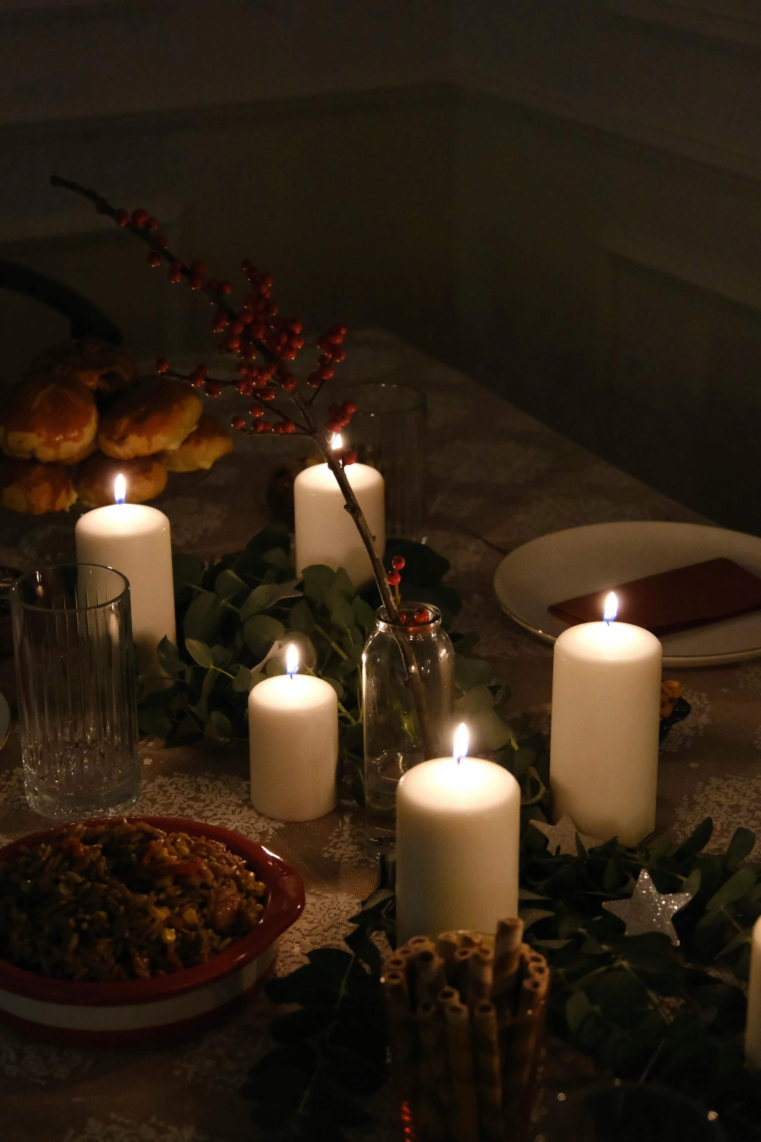 A table lit with candles, on which are placed dishes like breads and casseroles. A sprig of winter berries sits in a glass jar at the center.