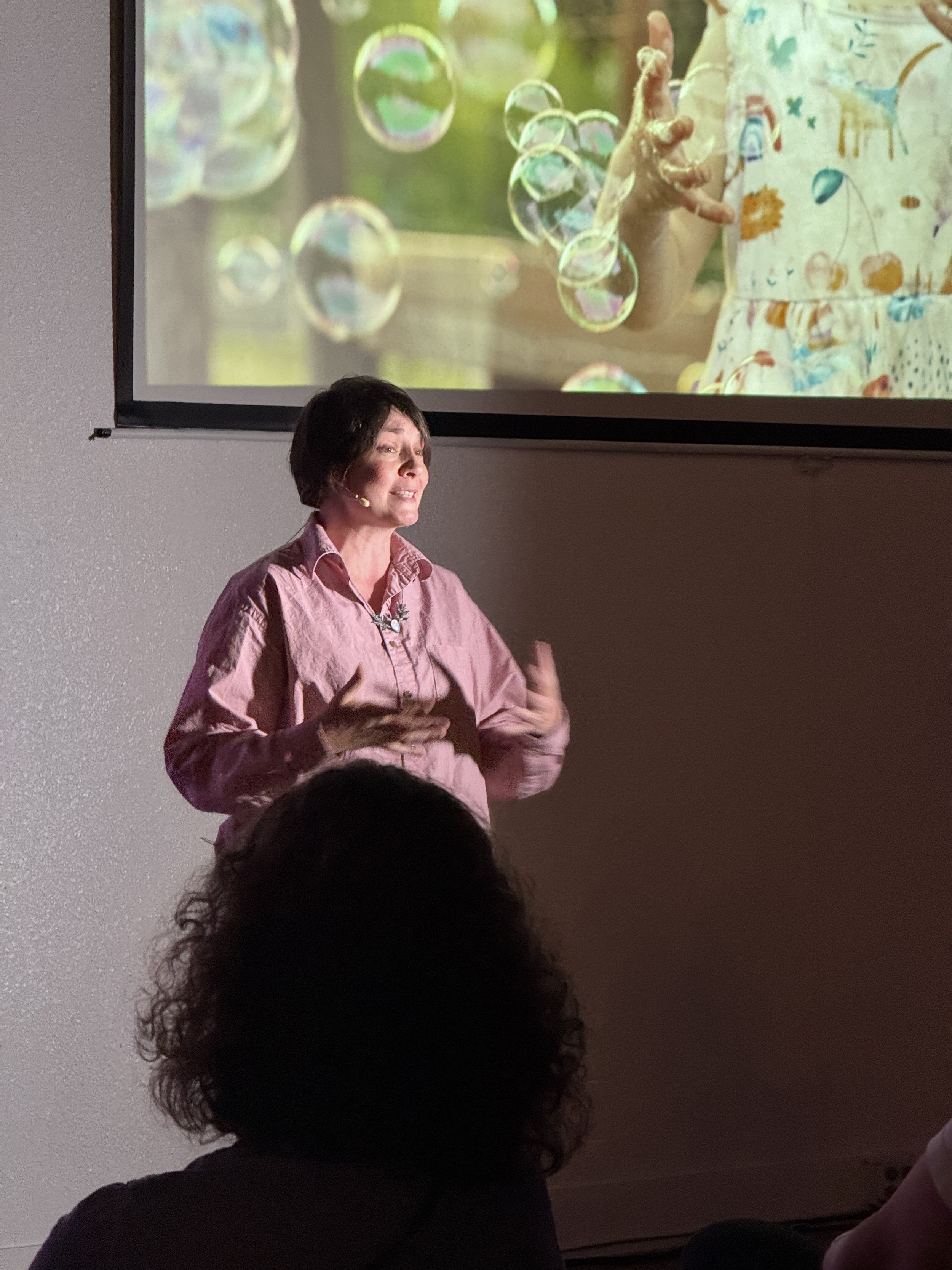 Ali McGhee, director of the Enneagram School of Awakening, giving a presentation in front of a projection of a child playing and reaching for soap bubbles outdoors. The woman wears a pink shirt and is speaking to an audience.