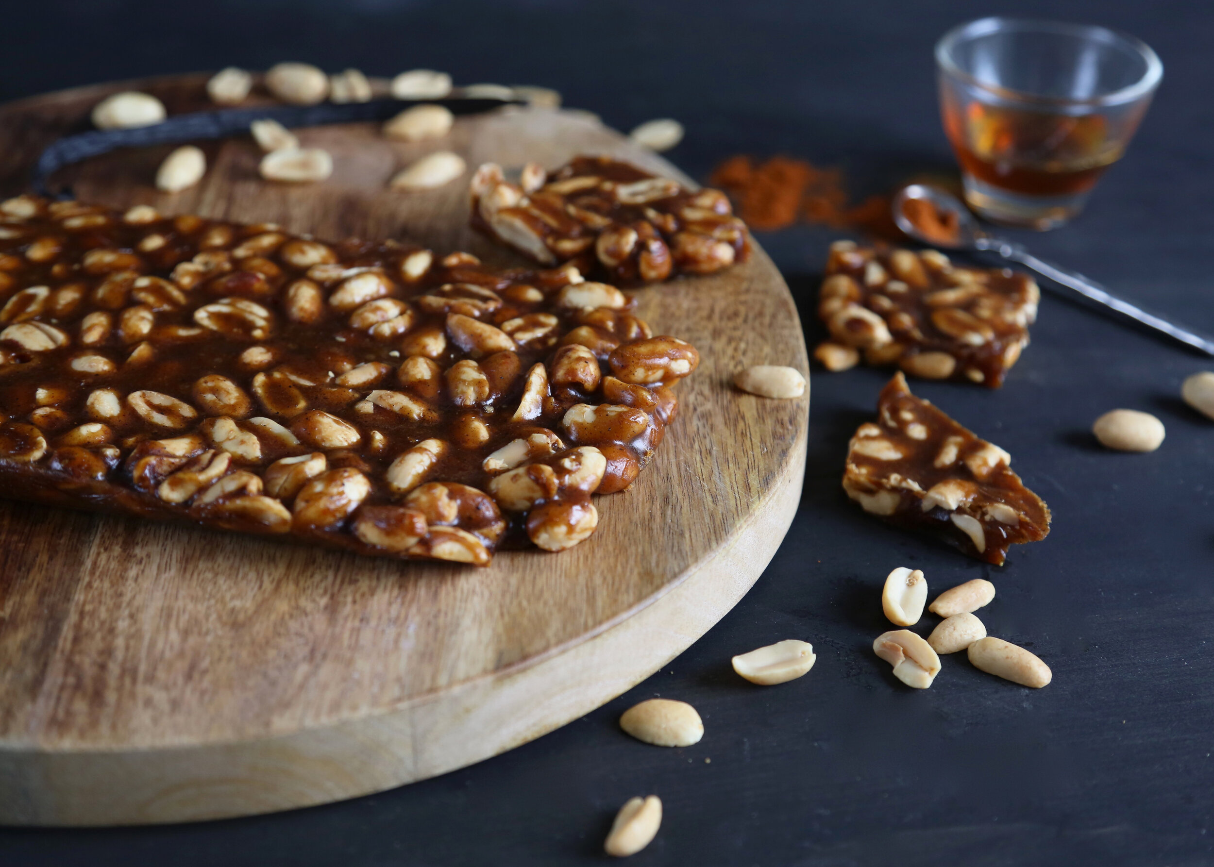 Homemade peanut brittle cut into pieces on a wooden board, with scattered peanuts and a glass of tea.
