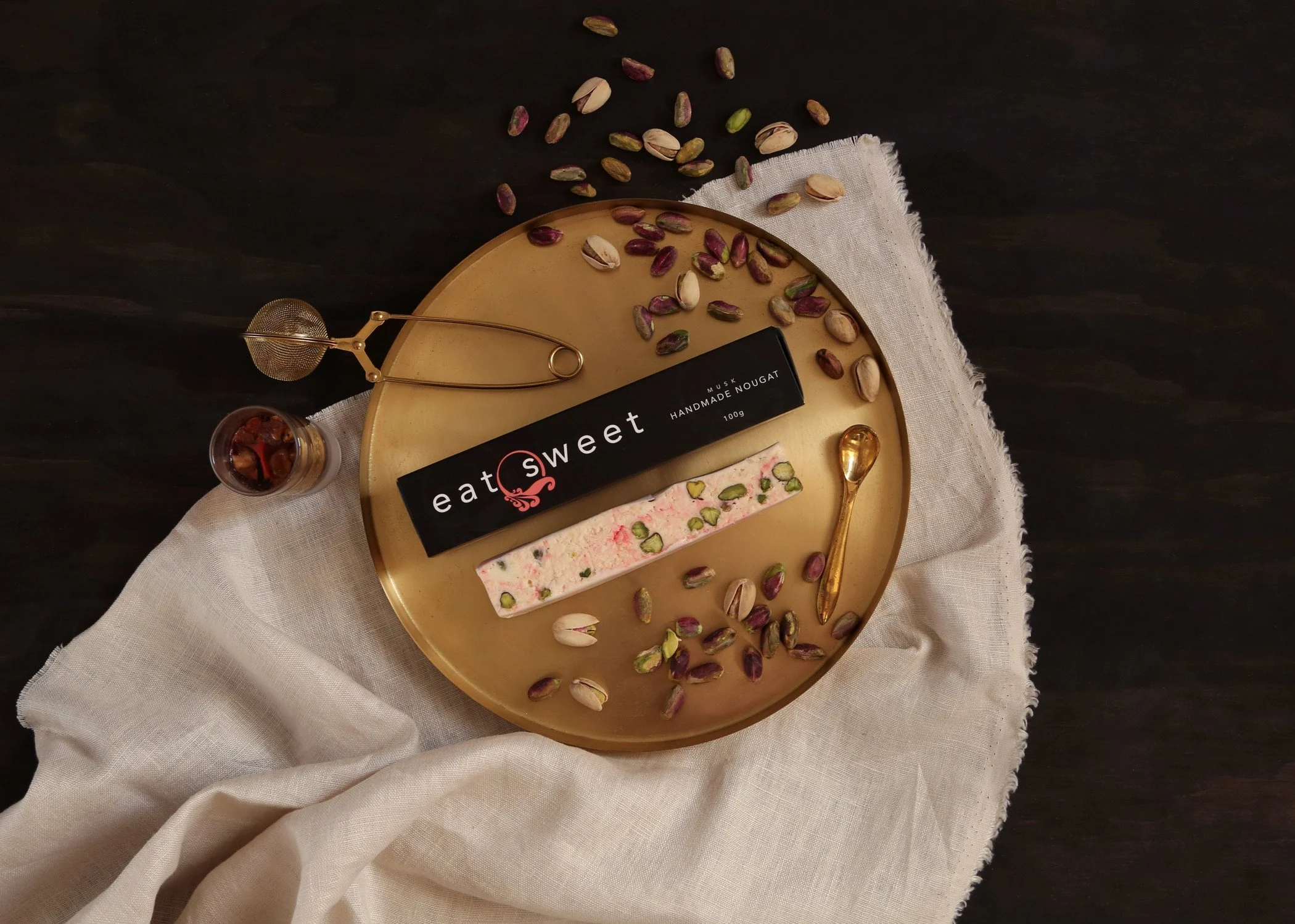 Overhead display of pistachio nougat on a gold tray with scattered pistachios, a tea infuser, spoon, and small jar on black wood.
