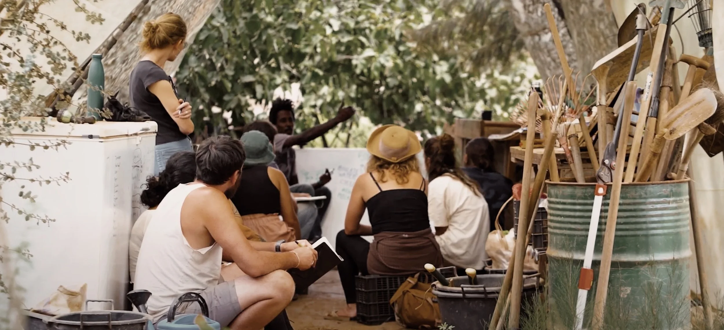 Students in sun hats sitting in the shade, watching facilitator Kyle demonstrate dryland strategies on a whiteboard next to garden tools.
