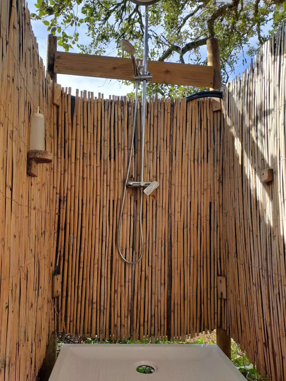 A rustic outdoor shower built from bamboo sticks, surrounded by vibrant green plants and garden foliage.