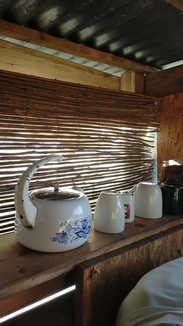 A tea pot and ceramic cups on a wooden shelf in the outdoor kitchen, with golden sunrays glowing in the background.