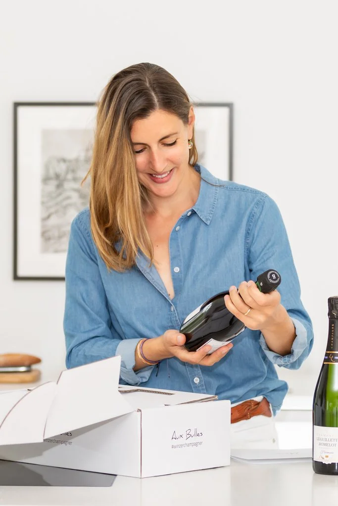 Eine junge Frau packt gerade eine Flasche Champagner aus einer Aux Bulles Champagnerabo Box und betrachtet sie lächelnd