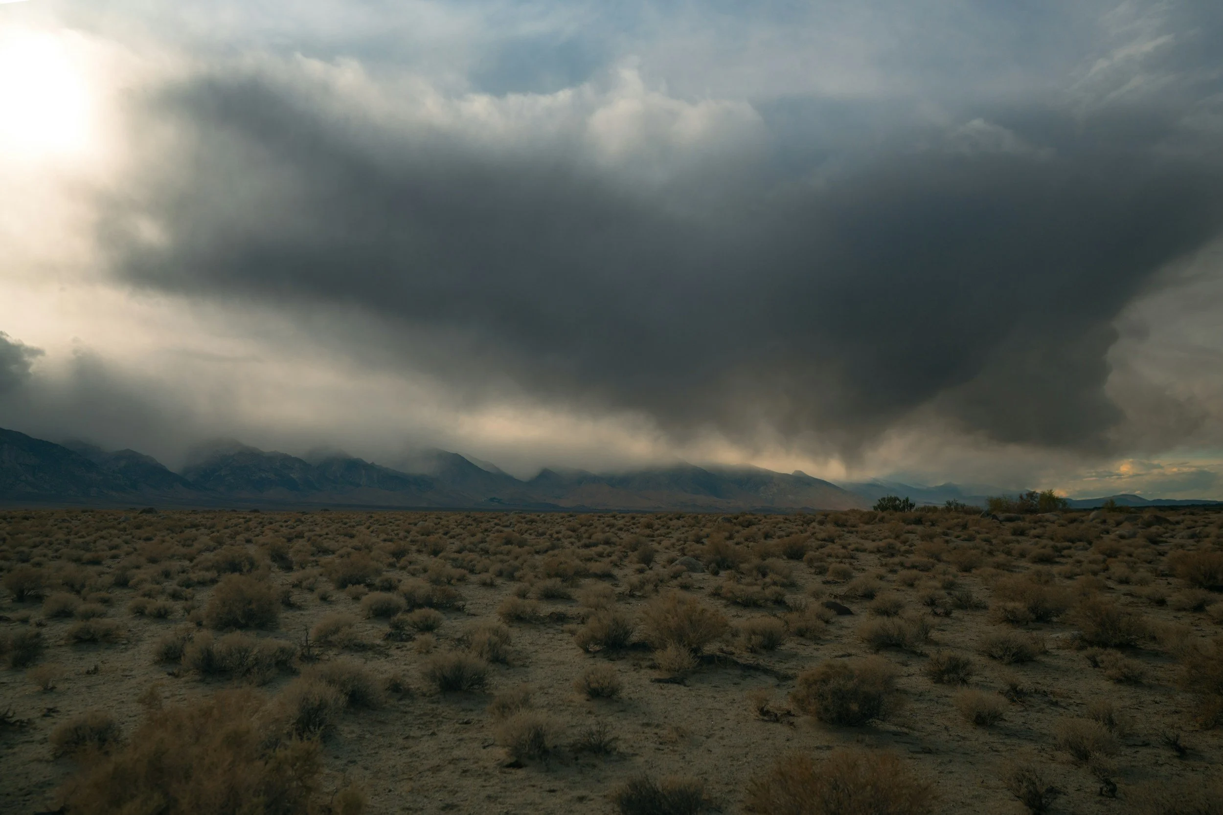 Storm clouds moving across a desert landscape with distant mountains and dry brush in muted evening light.