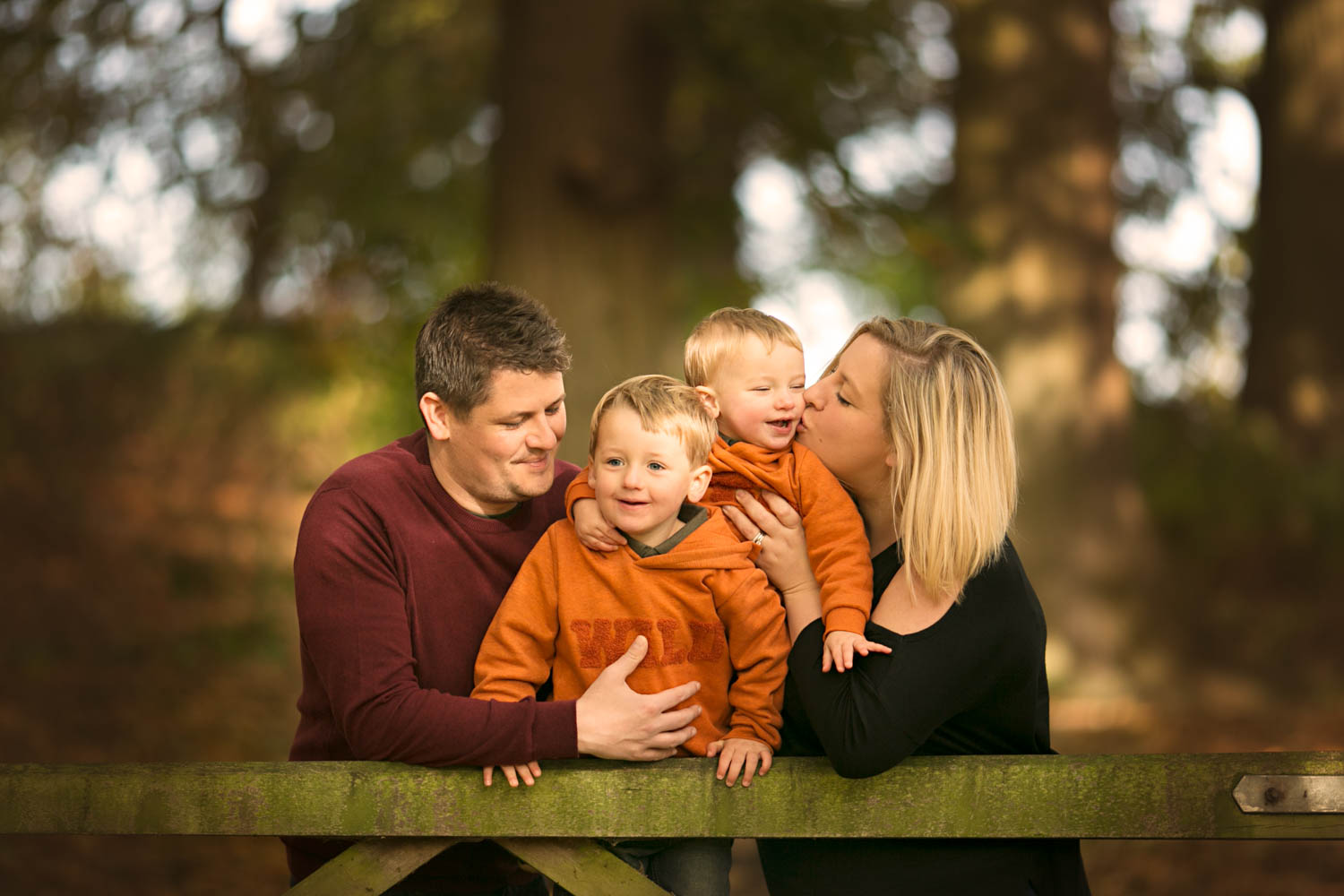  Family posing againgst a gate with mum kissing son during an Autumn family portrait session at Ampthill Park in Bedford. 