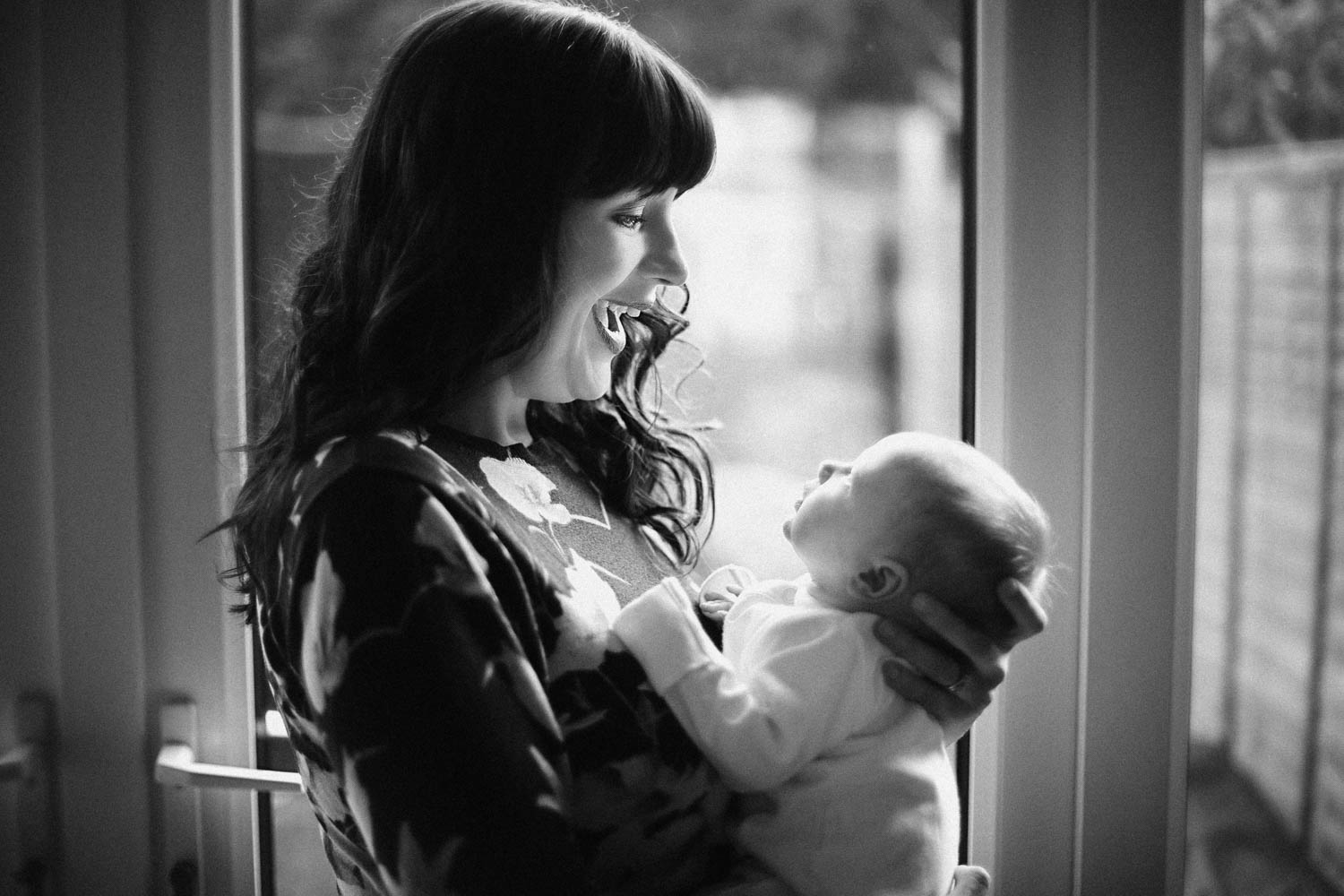  Black and White image of Mother and Newborn naturally posing in front of window during baby lifestyle photoshoot at clients home in Bedford. 