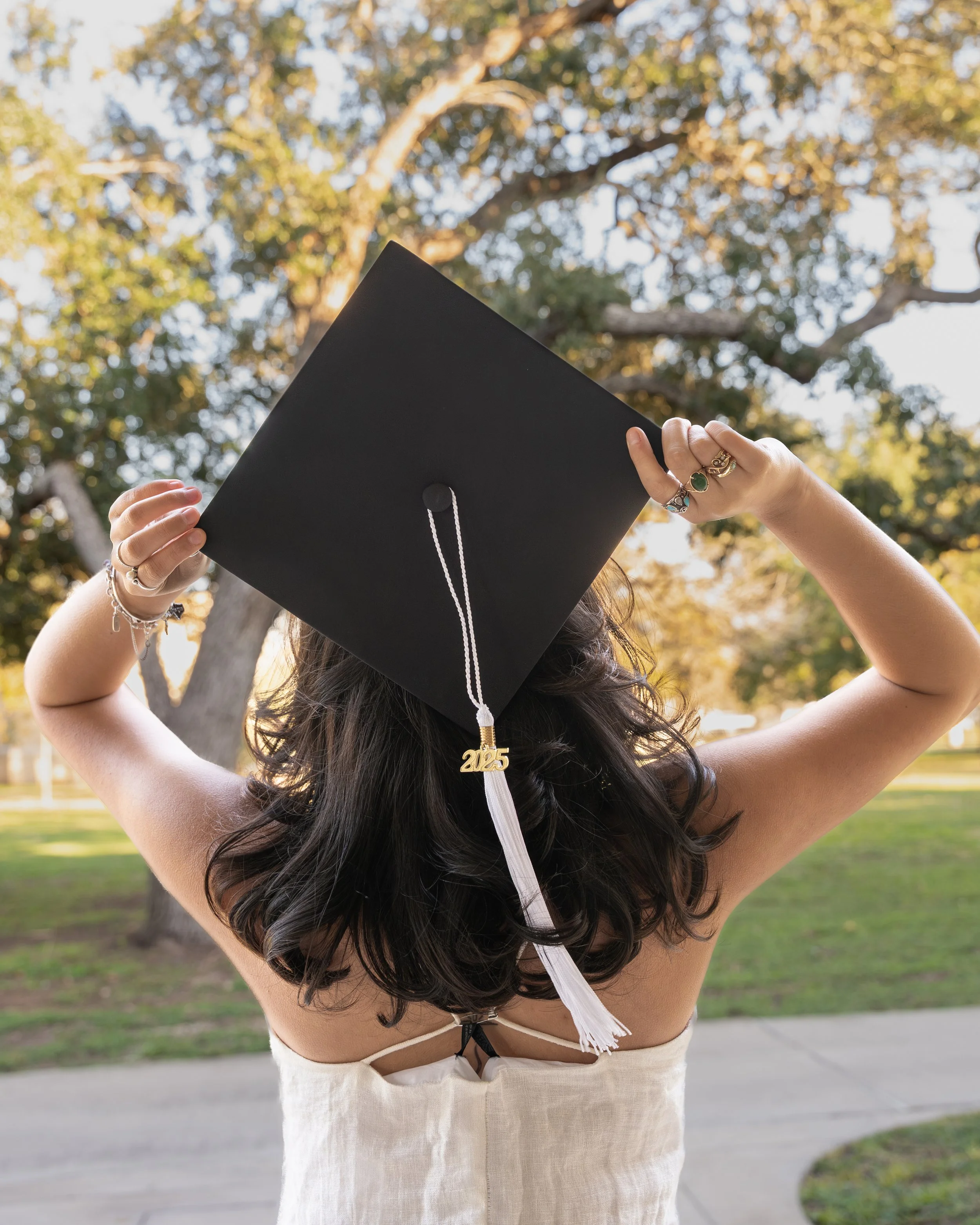 Person holding a black graduation cap with a white tassel and a gold '2025' charm, outdoors with trees and clear sky in the background.