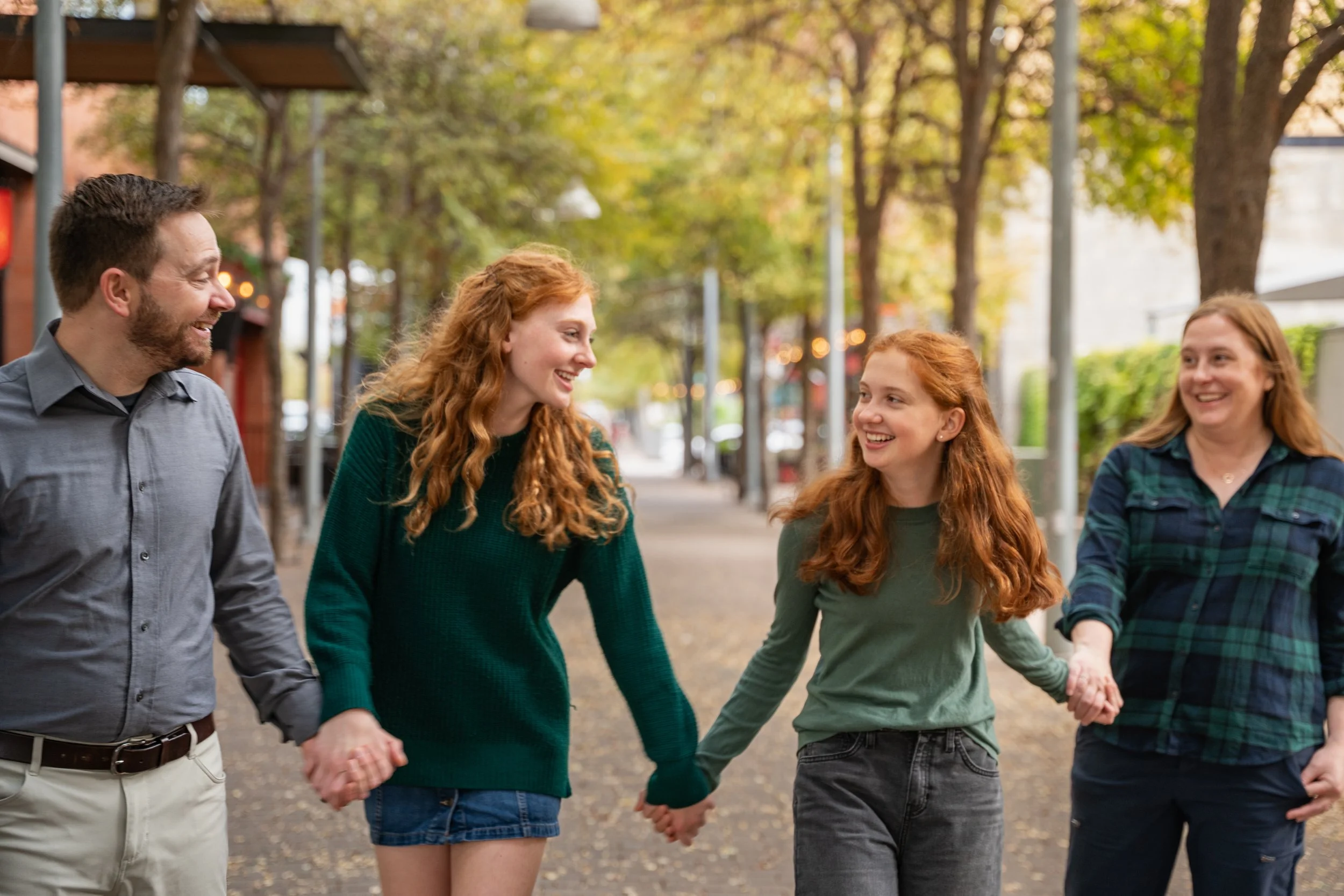 A group of four people, two men and two women, holding hands and walking outdoors on a fall day. They are smiling and engaging with each other, surrounded by trees with yellowing leaves.