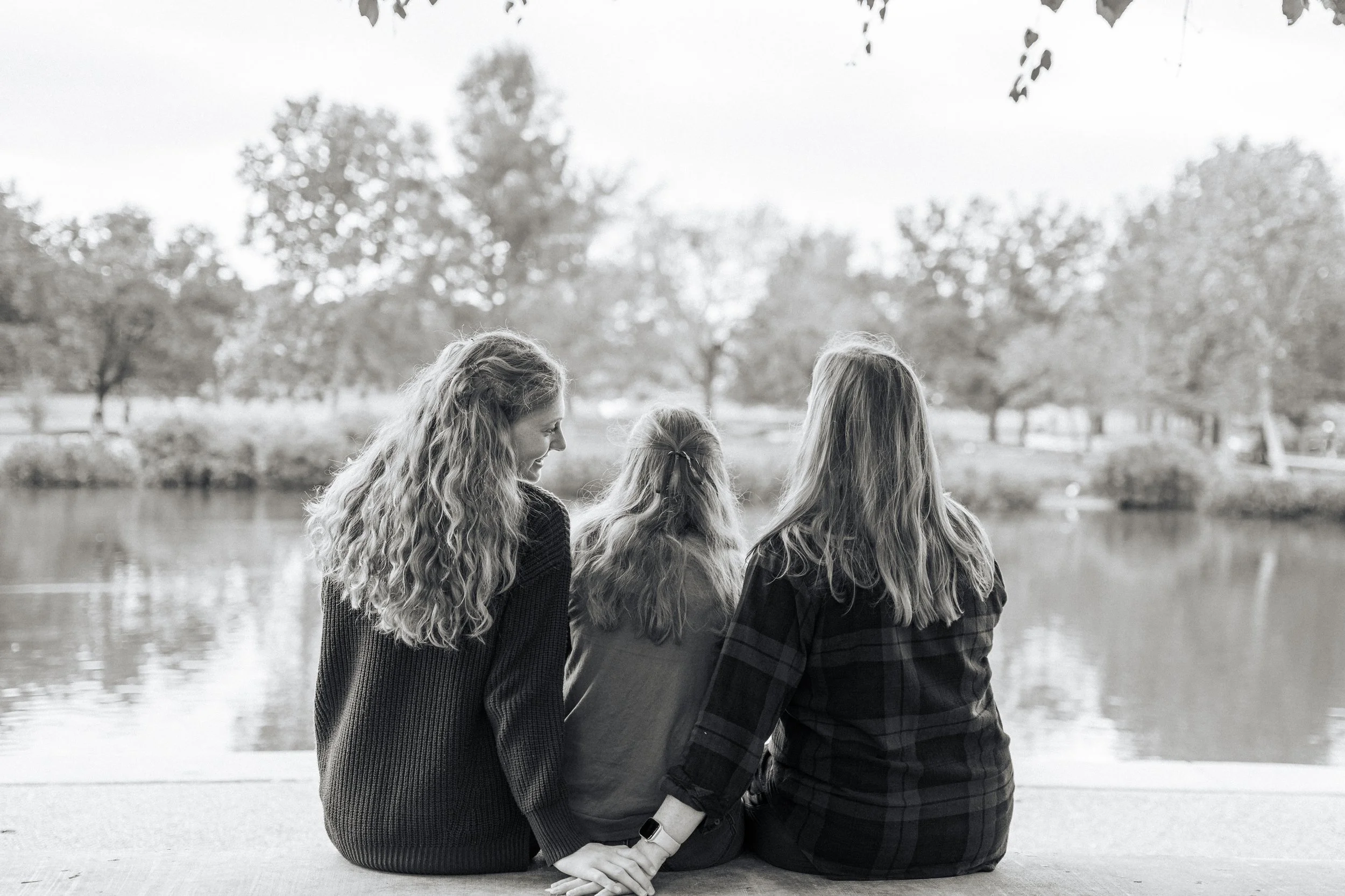 Four women with long hair sitting by a lake, holding hands, with trees in the background.