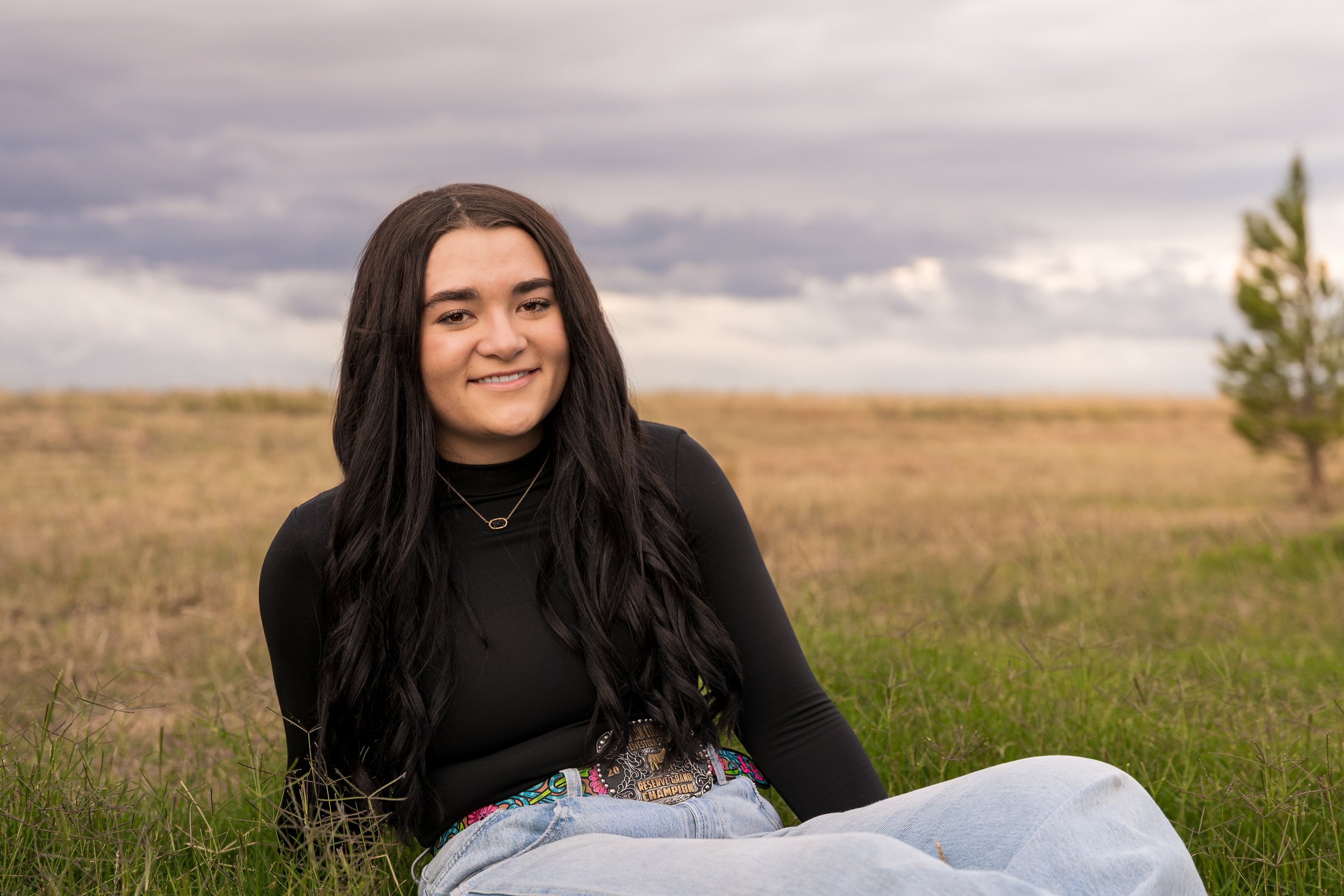 A young woman with long dark hair smiling while sitting on a grassy field under a cloudy sky, wearing a black top, light blue jeans, and a championship belt.