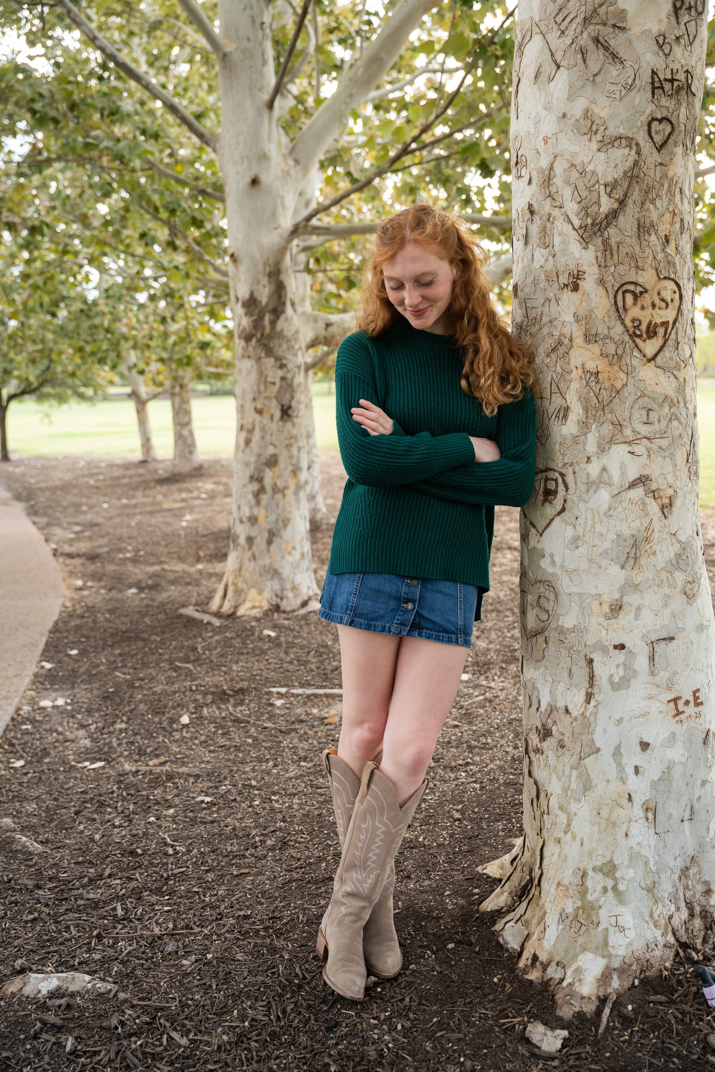 A young woman with long curly red hair leaning against a tree trunk in a park, wearing a dark green sweater, a denim skirt, and cowboy boots. The tree has carved initials inside a heart on its bark. The background features other trees and a grassy area.