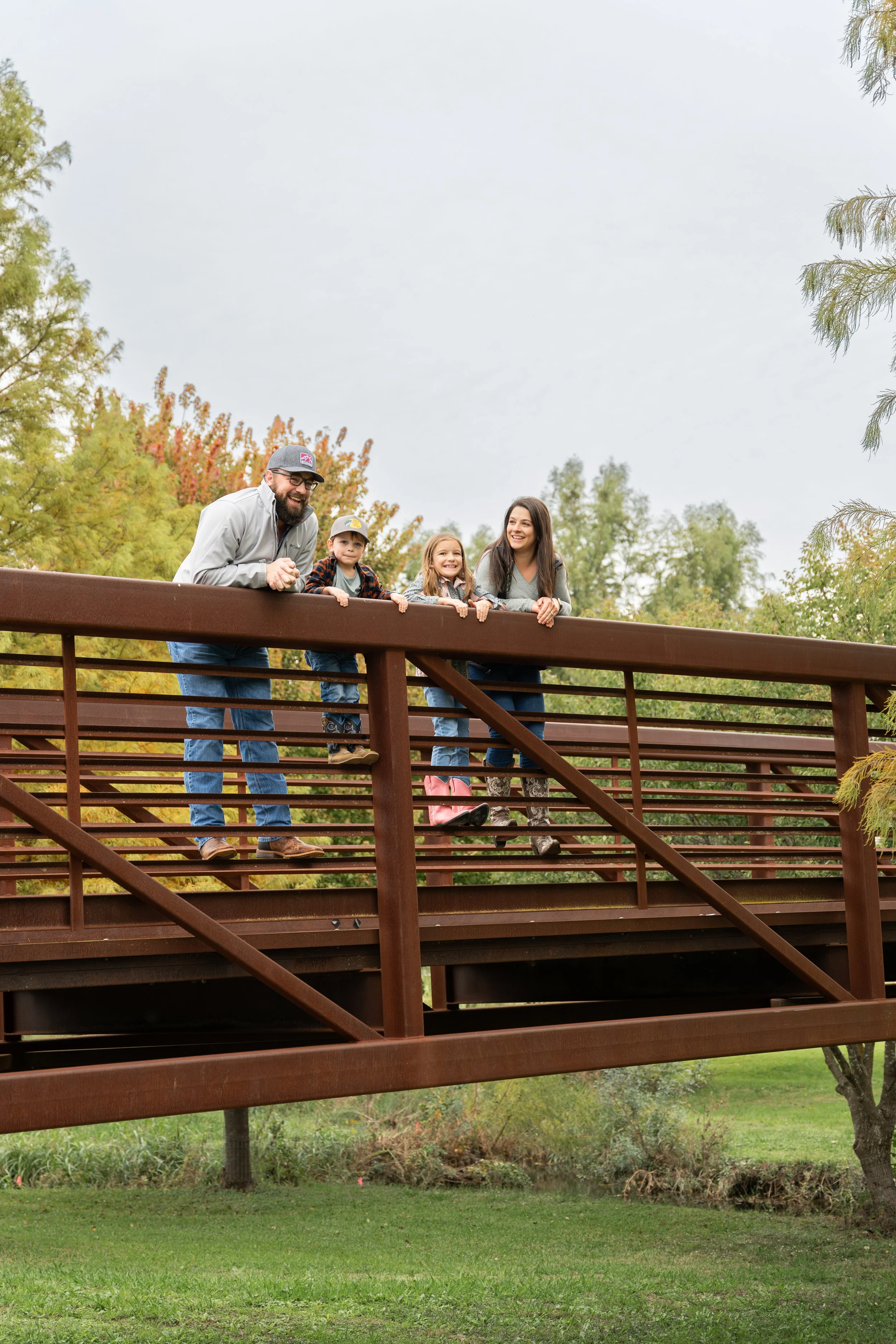 Four people, including two children and two adults, leaning on a brown wooden bridge railing, smiling and looking at each other, with trees and grass in the background.