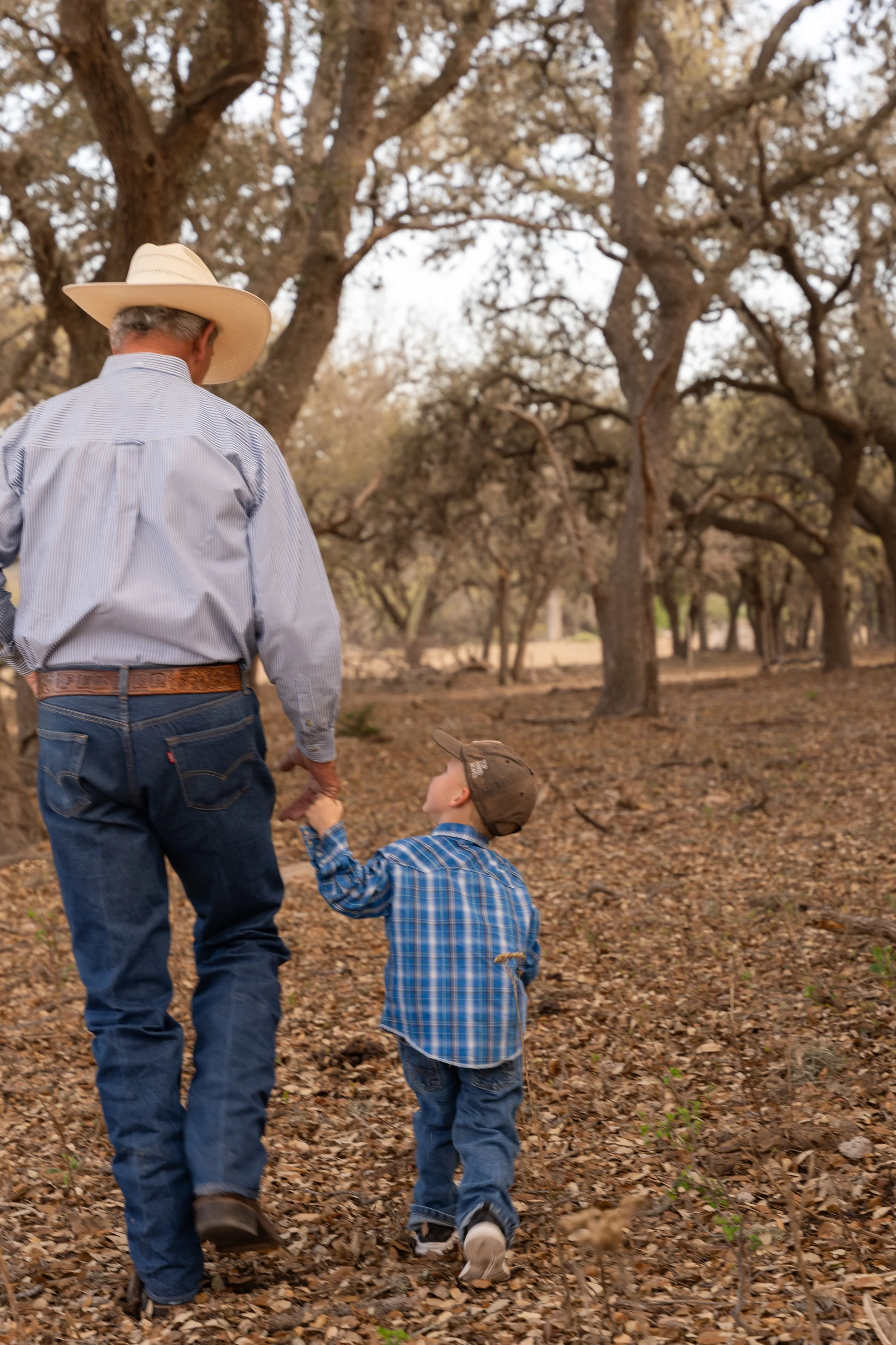 A man wearing a cowboy hat and a striped shirt walking with a young boy in a plaid shirt and cap through a wooded area with many large leafless trees.