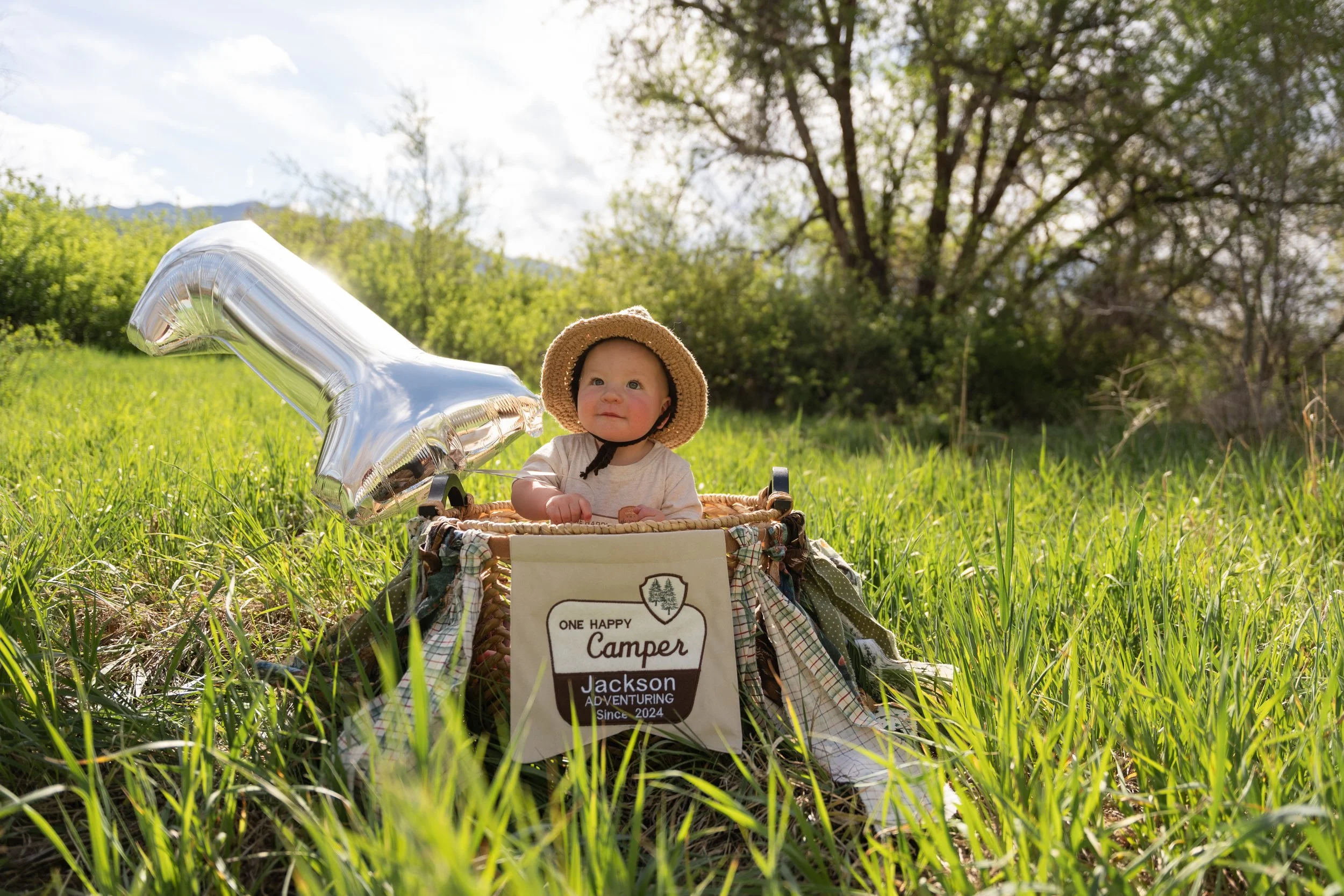 A baby wearing a sun hat sits in a basket in a grassy field holding a small toy, with a large silver balloon in the shape of the number '1' beside them. There is a sign in the basket that reads 'ONE HAPPY CAMPER Jackson ADVENTURING Since 2024'. In the background, there are trees and a partly cloudy sky.