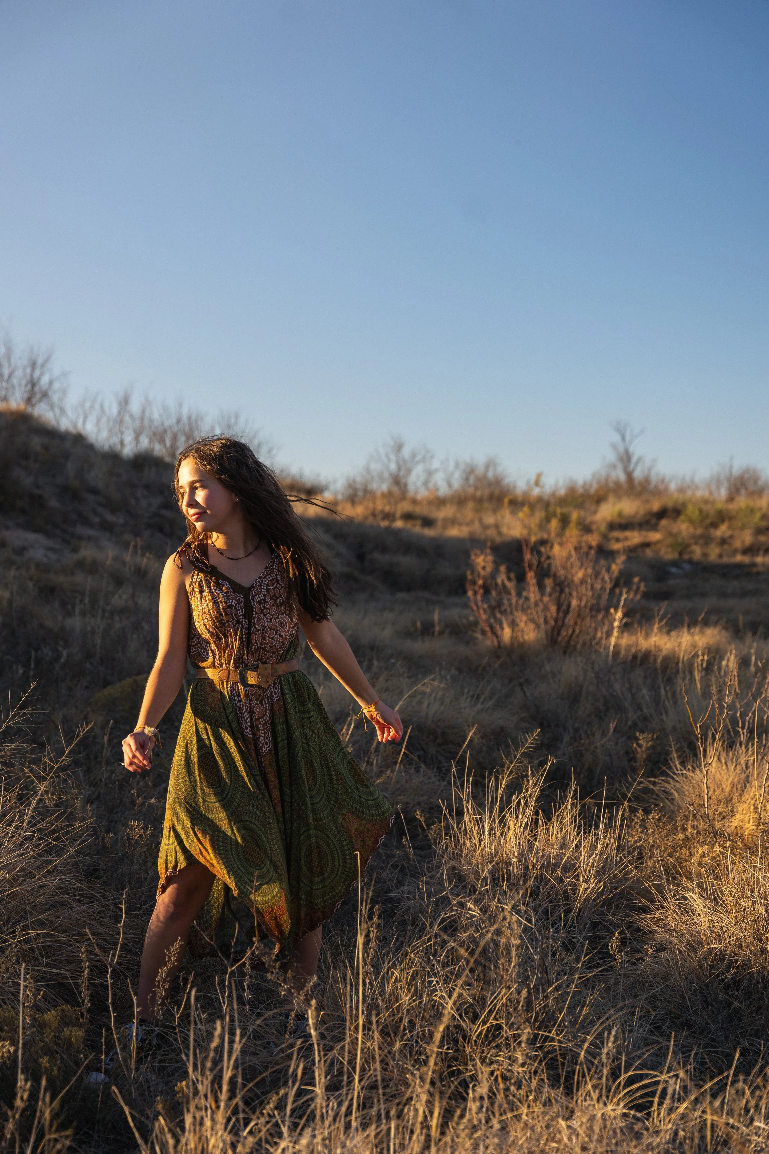 A young woman with long wavy hair and a patterned dress walking through tall dry grass in an open, sunny landscape with sparse bushes and clear sky.
