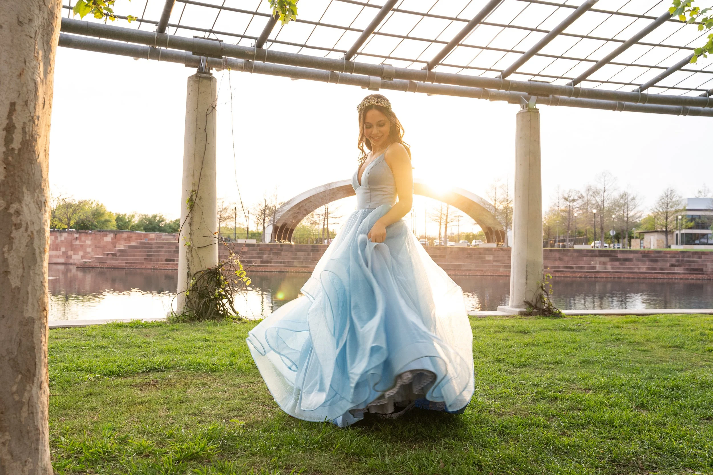A woman dressed as a princess in a light blue gown, wearing a tiara, is standing outdoors by a river, smiling as sunlight shines behind her. There are trees, a stone arch, and a bridge in the background.