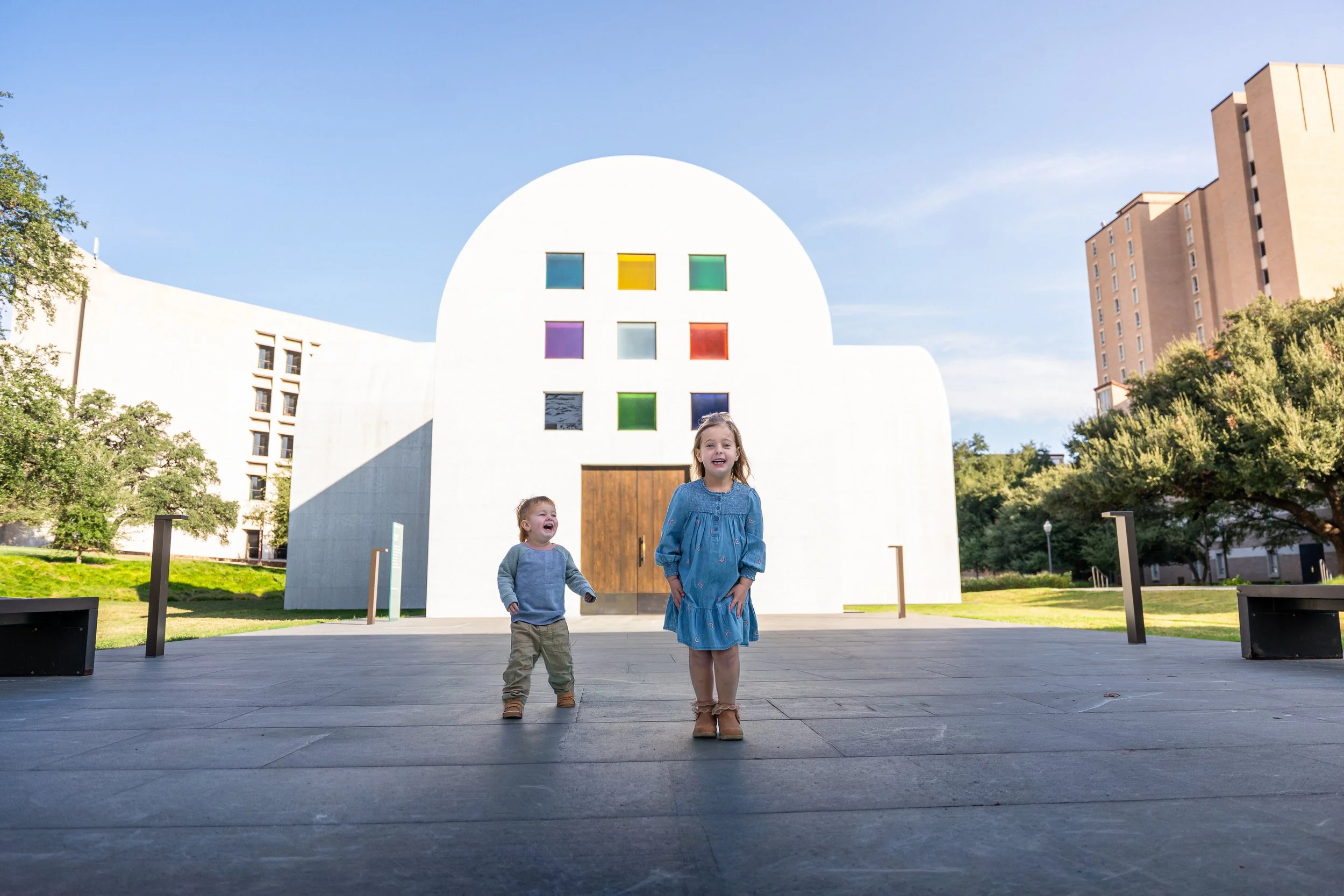 Two children, a girl and a boy, are walking on a paved area in front of a white modern building with colorful square windows. The girl is wearing a blue dress and smiling, while the boy is wearing a gray shirt and beige pants, looking happy. The scene is outdoors with trees and tall buildings in the background, during daytime with a clear blue sky.
