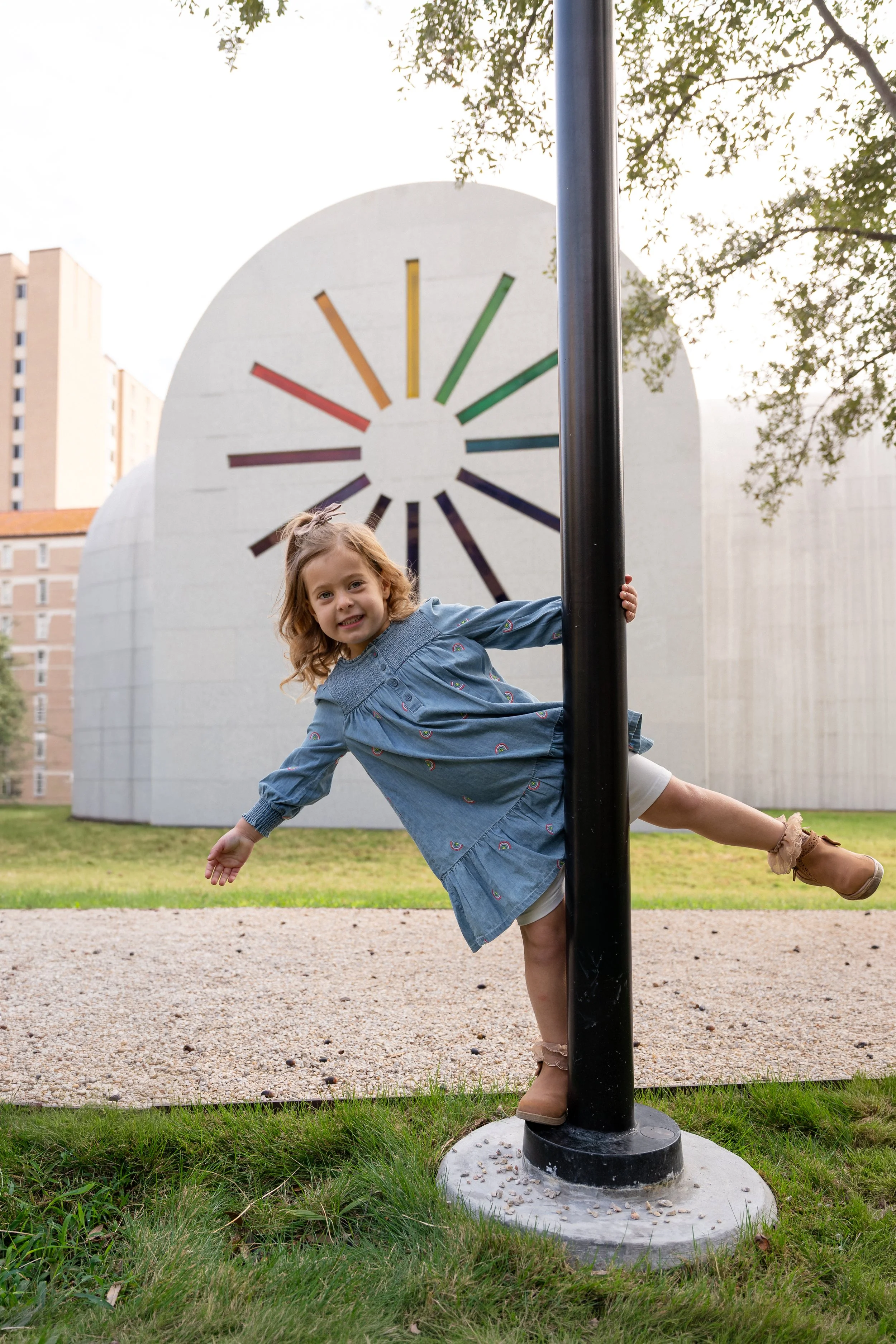 A young girl with curly blonde hair is smiling and playing on a playground, hanging onto a black pole with one hand and leaning to the side with one leg in the air. She is wearing a blue dress with rainbow patterns and tan shoes. In the background, there is a large white building with a colorful circular design, trees, and taller residential buildings.