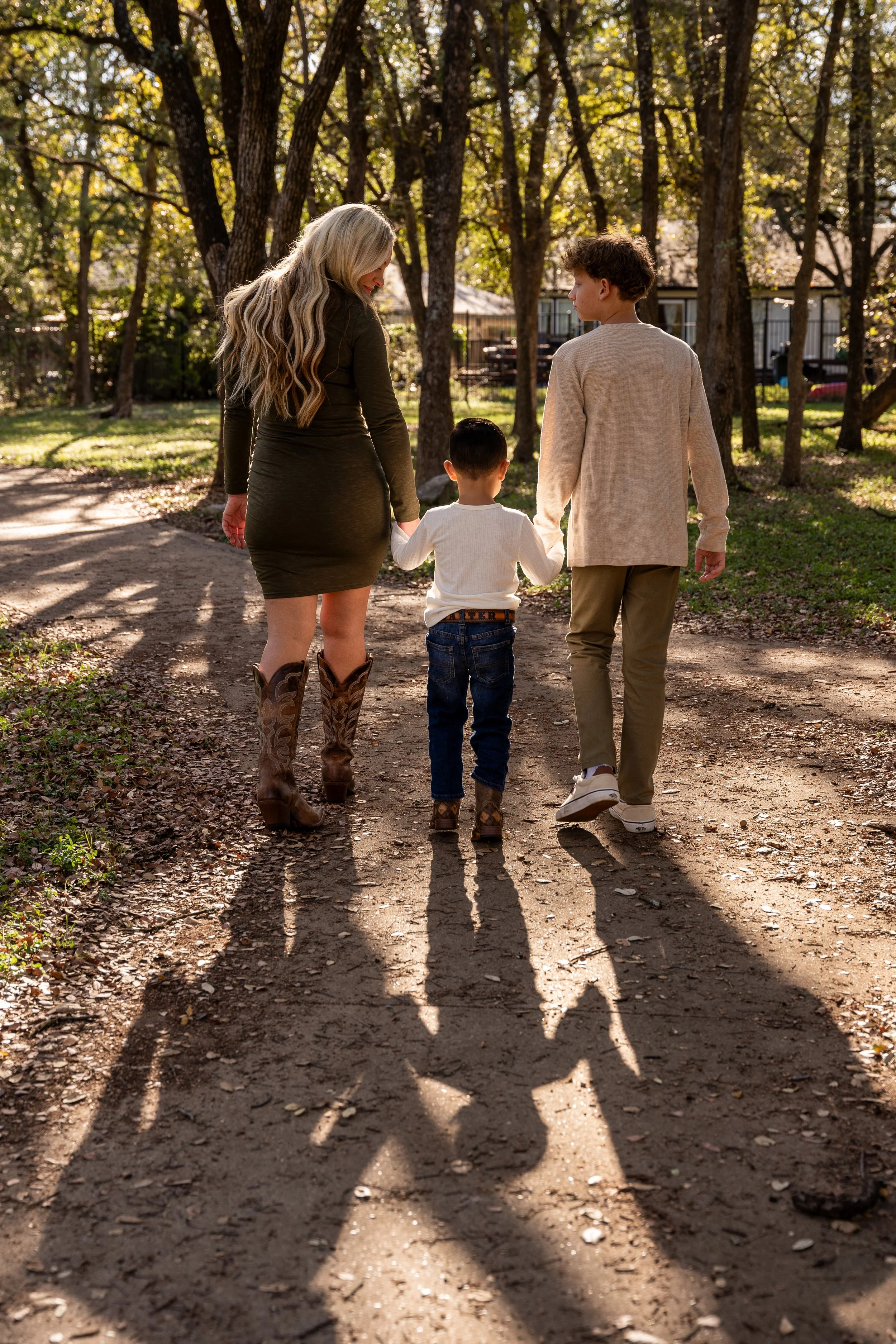 A woman and two children walking hand in hand on a dirt path surrounded by trees and sunlight, casting long shadows.