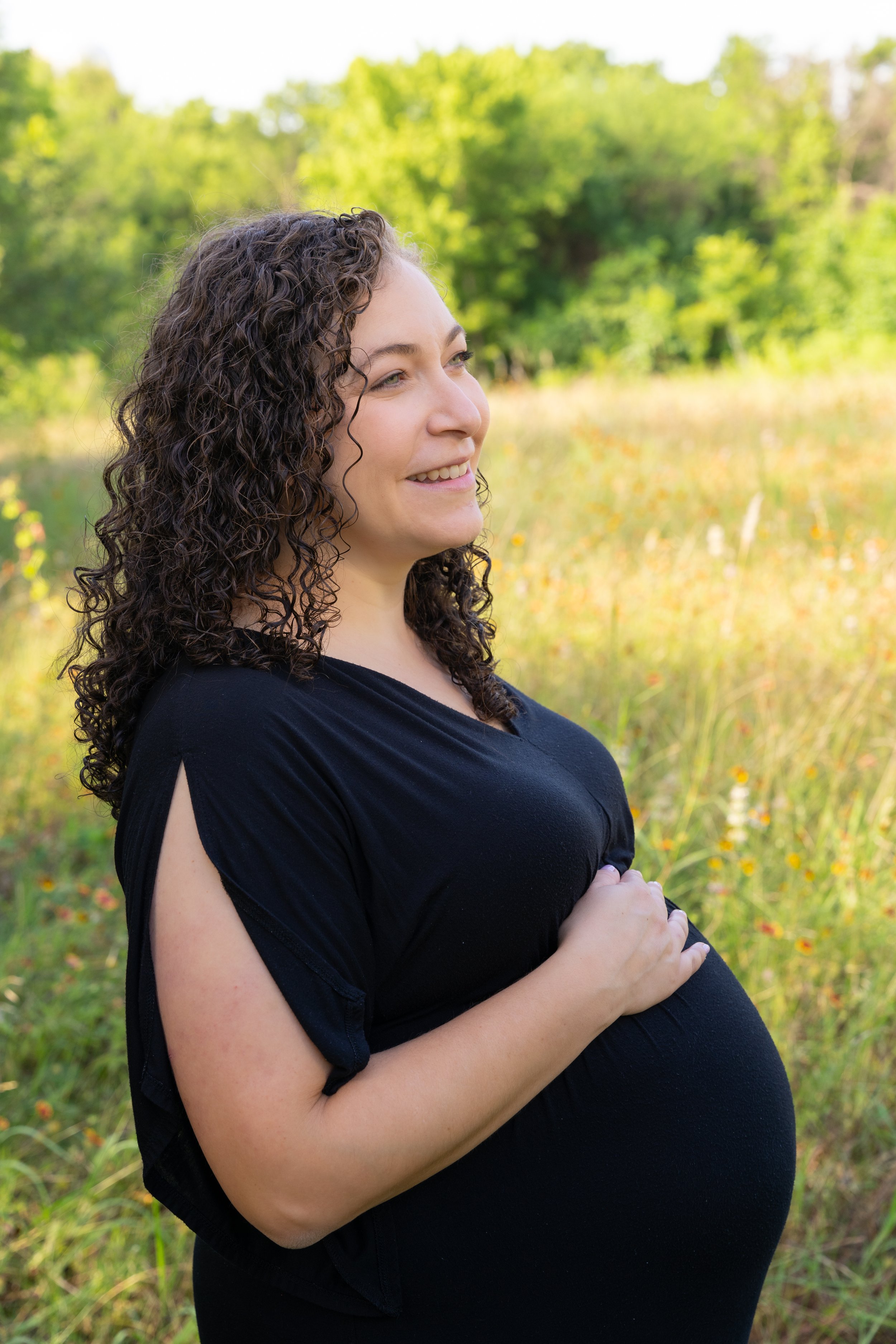 A pregnant woman with curly hair standing outdoors in a field with green trees in the background, smiling and looking to her left.