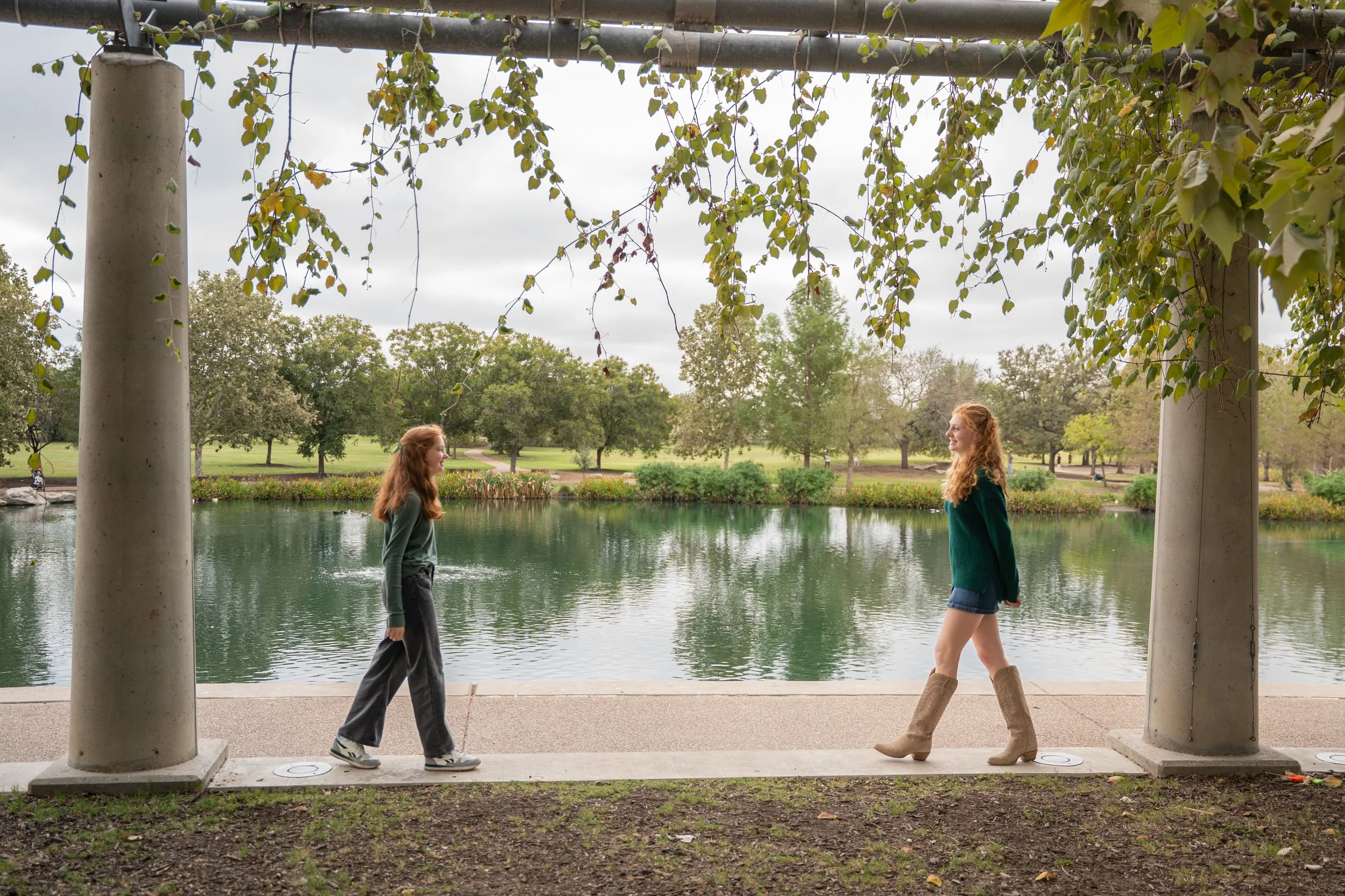 Two women with red hair are standing on a sidewalk under a pergola with hanging vine plants, near a lake in a park. They are facing each other, smiling, and wearing casual clothing; one has on boots, the other sneakers.