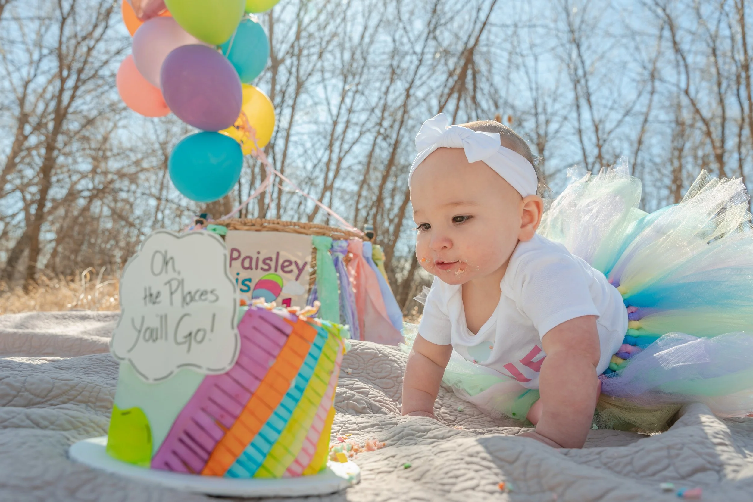 A baby dressed as a fairy at an outdoor birthday celebration, with colorful balloons and a decorated birthday cake in the foreground.