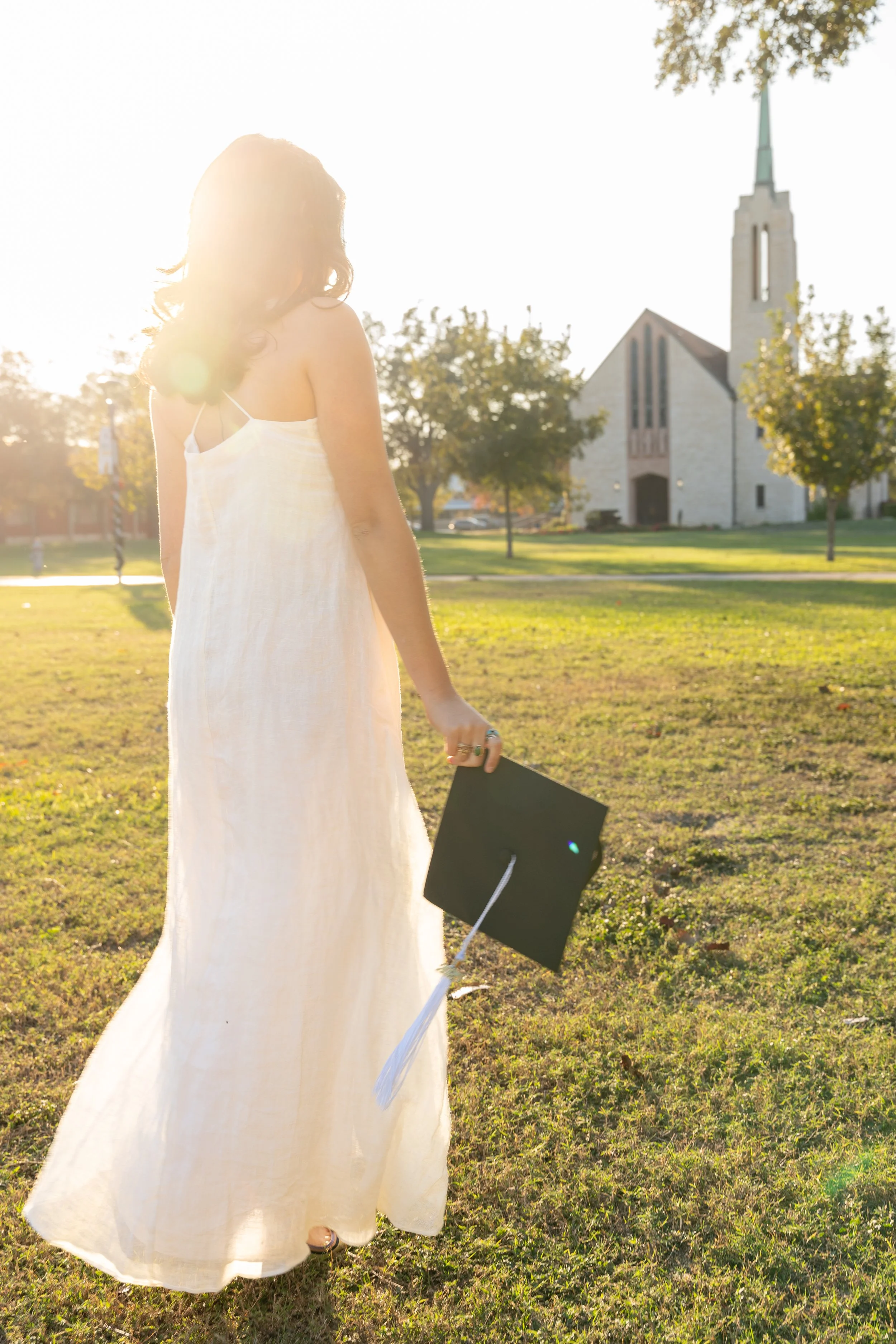 A woman in a white dress holding a black graduation cap, standing on a grassy field near a church, with the setting sun behind her.