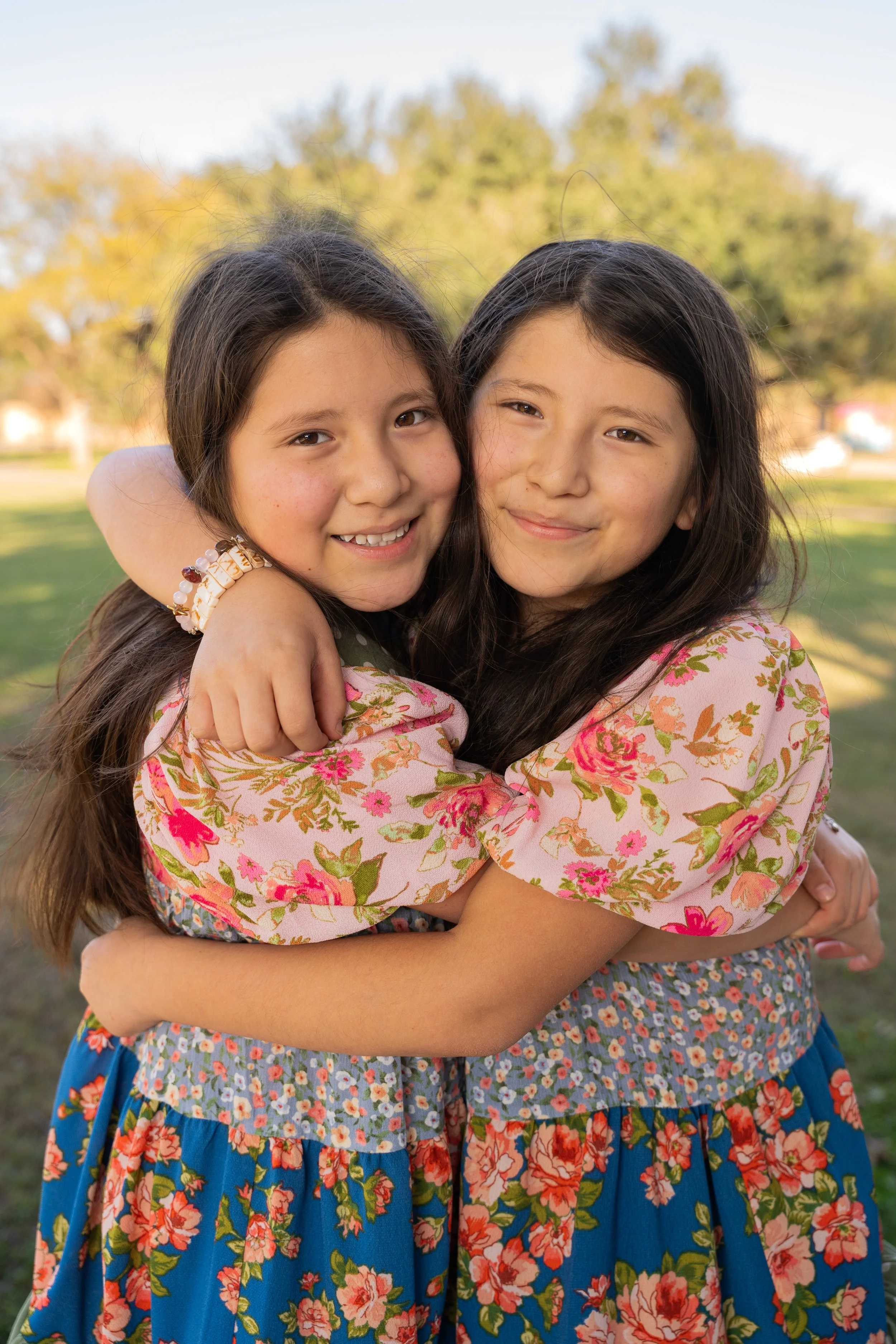 Two young girls hugging and smiling outdoors, wearing floral-patterned dresses, with a blurred background of trees and sky.