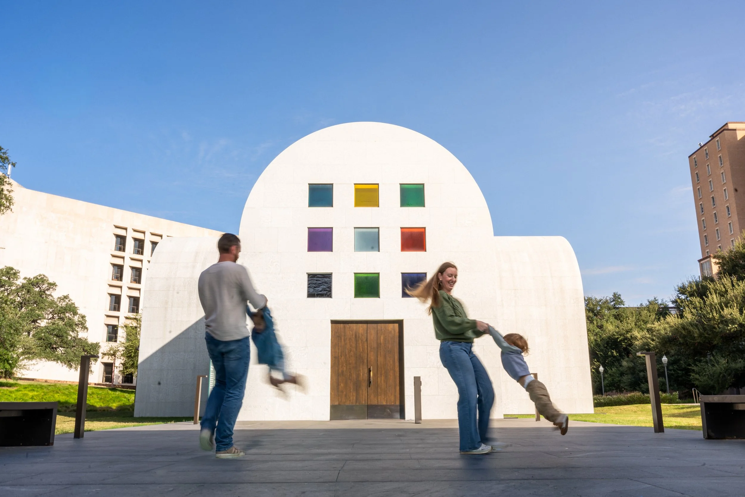 People playing and spinning around in front of a modern white building with colorful square windows on a sunny day.