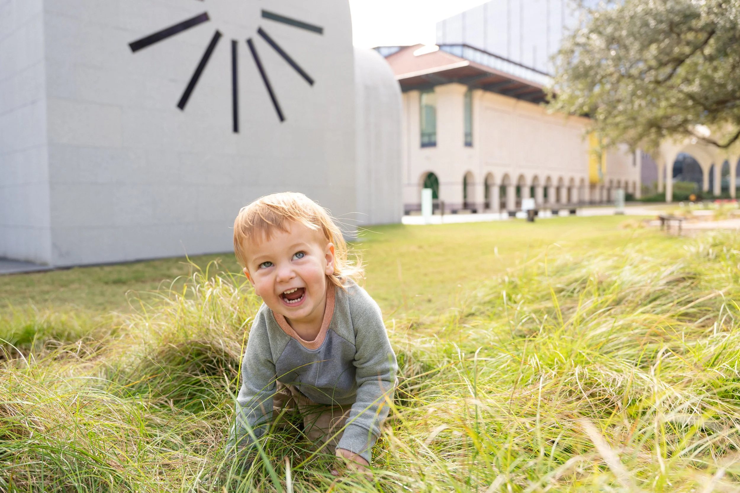 A young child with blonde hair and blue eyes crawling on a grassy area with buildings and trees in the background.
