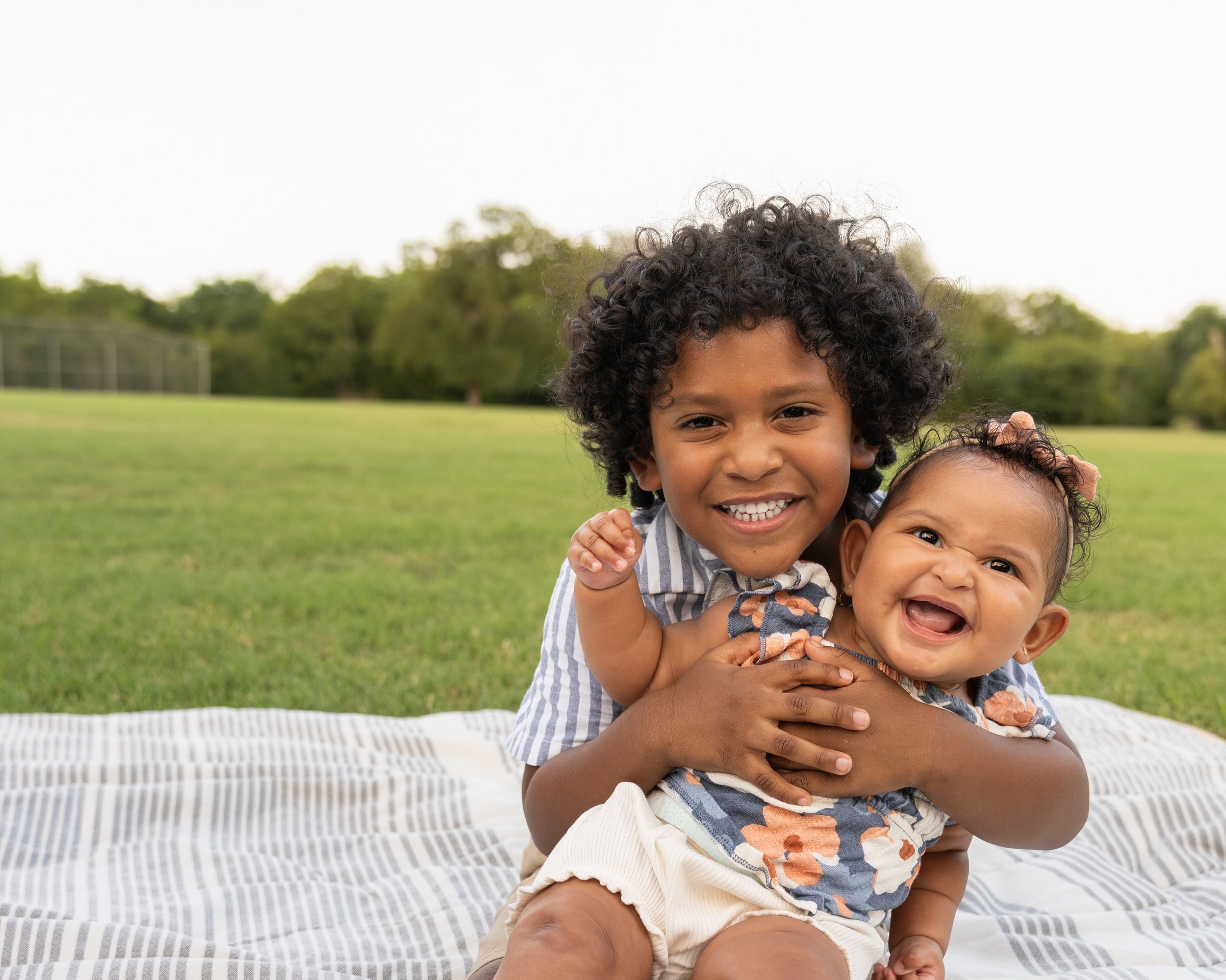Young boy with curly hair smiling and hugging a younger girl outdoors on grass.