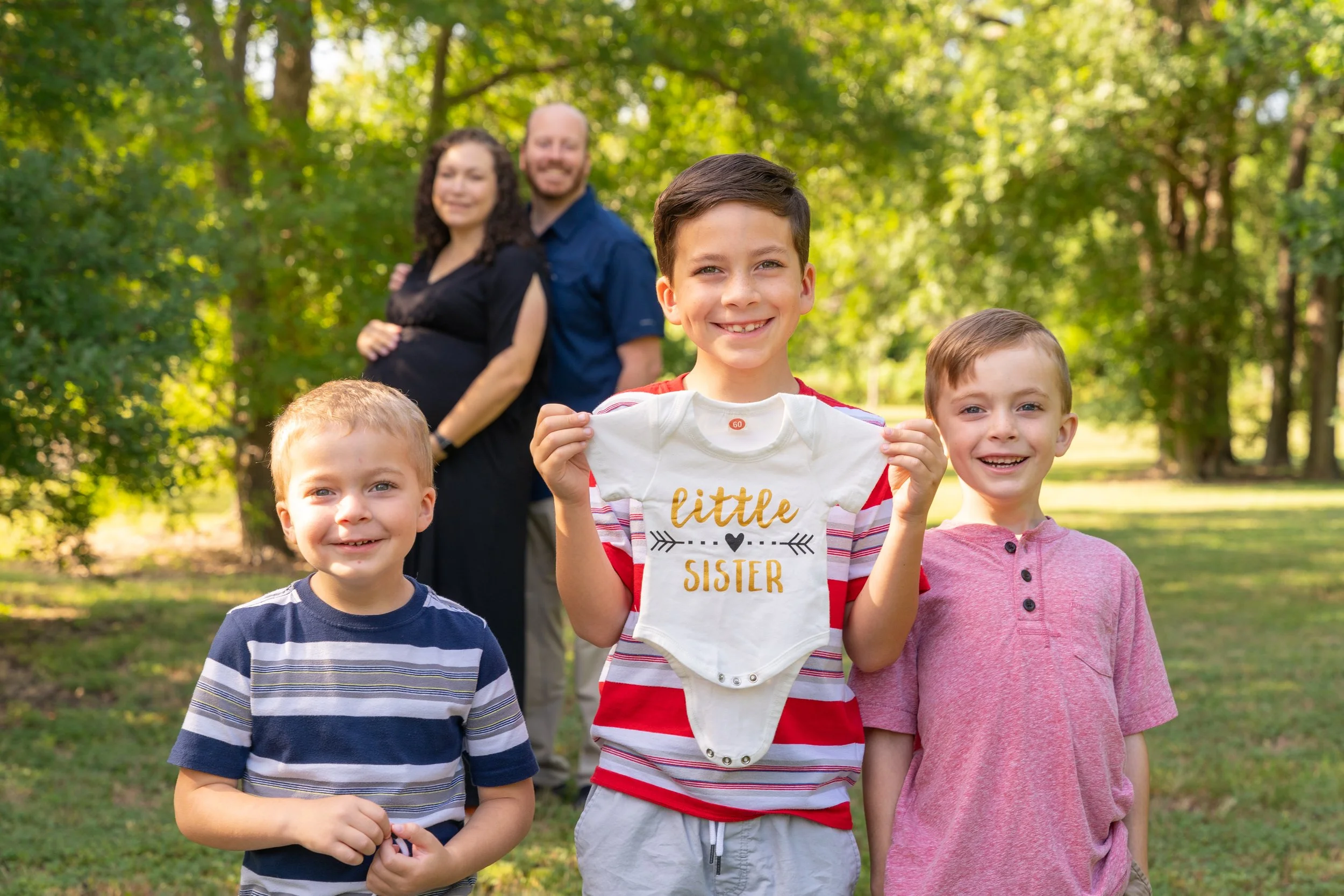 A group of smiling children standing outdoors with a family of three in the background, surrounded by green trees.