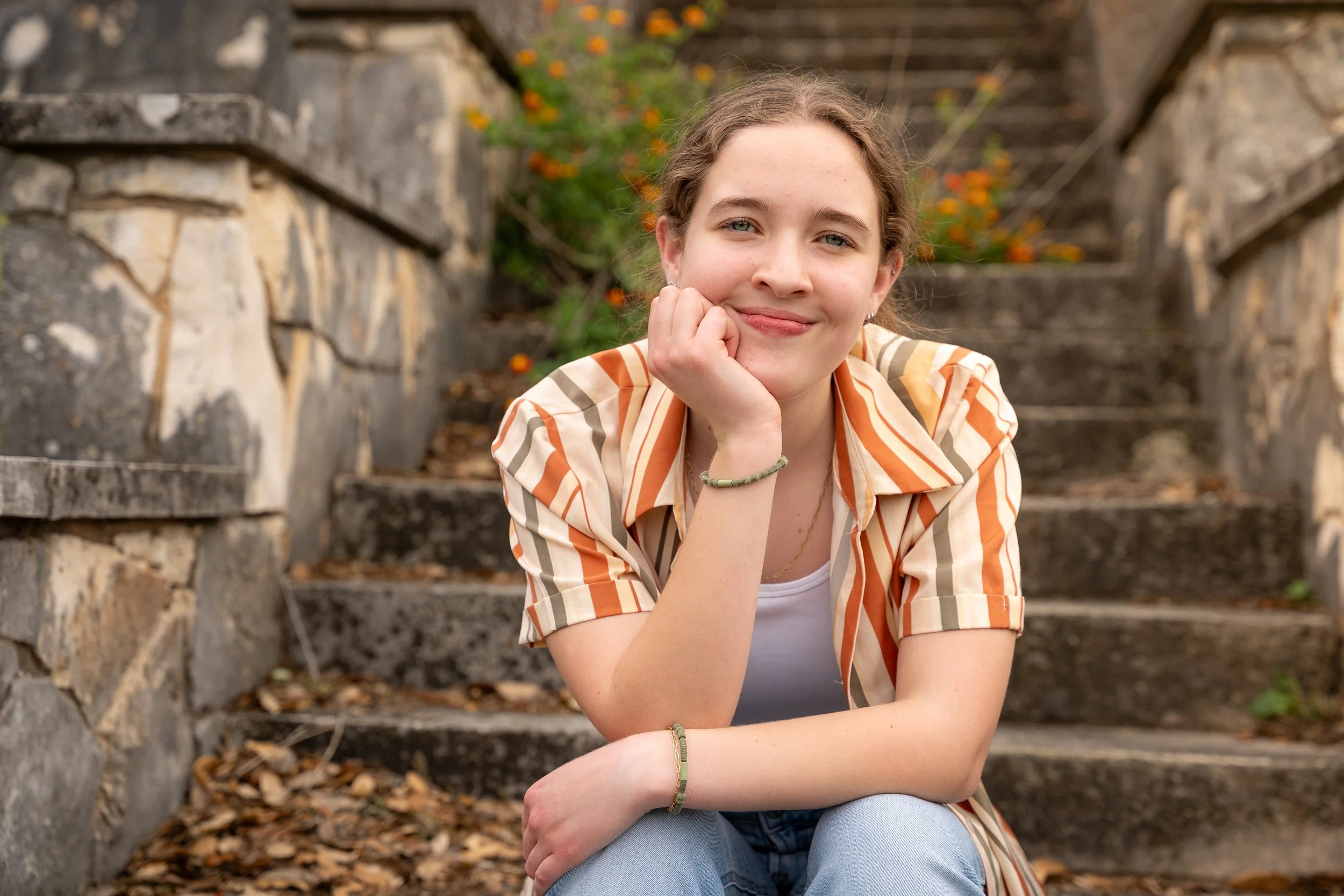 A young woman with curly light brown hair smiling, sitting on outdoor stone steps with fallen leaves, wearing a striped orange and beige shirt, a white top, and light blue jeans, with blurred plants and trees in the background.