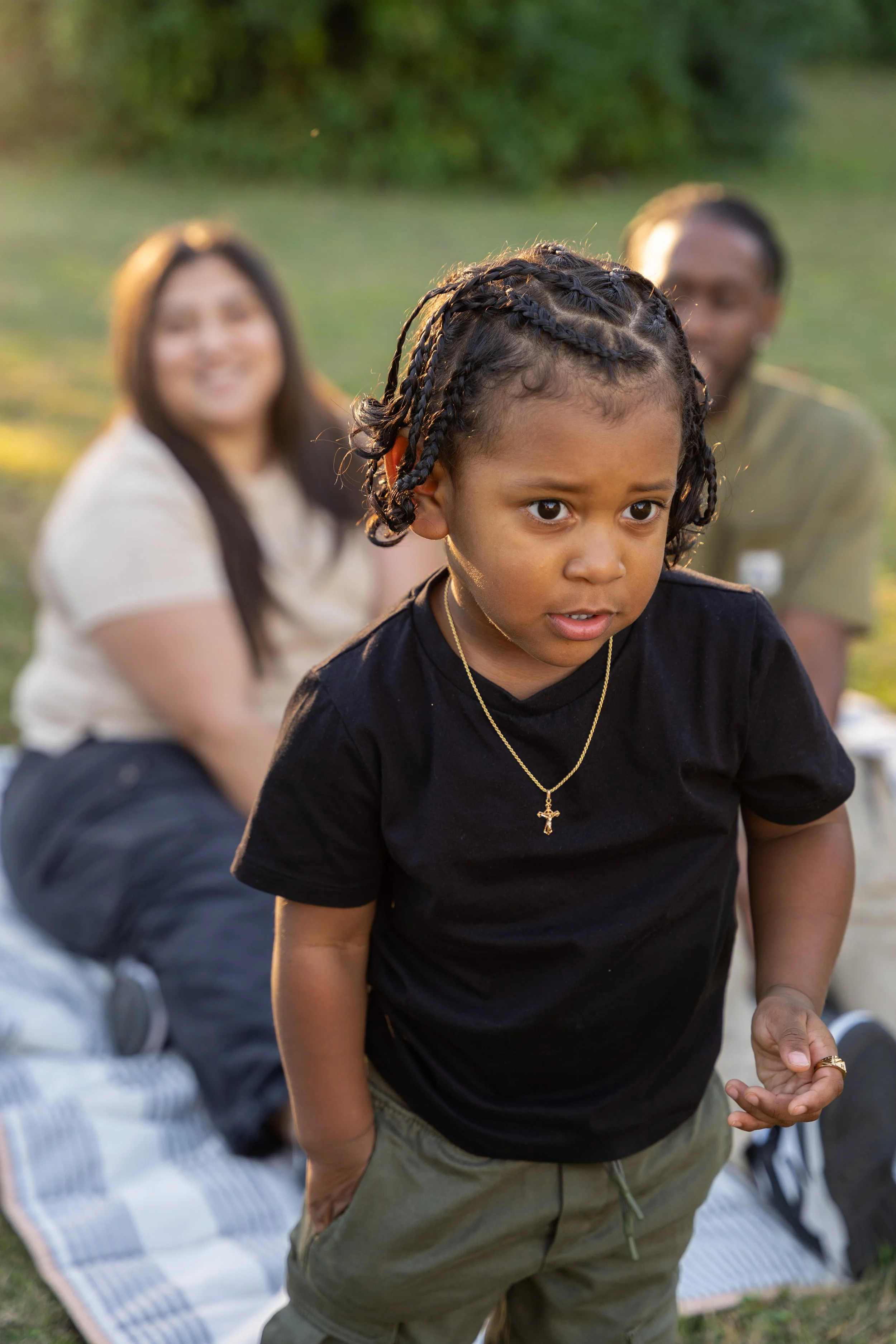 A young girl with braided hair and a cross necklace, standing outdoors with a blurry group of people behind her, on a grassy area during the day.