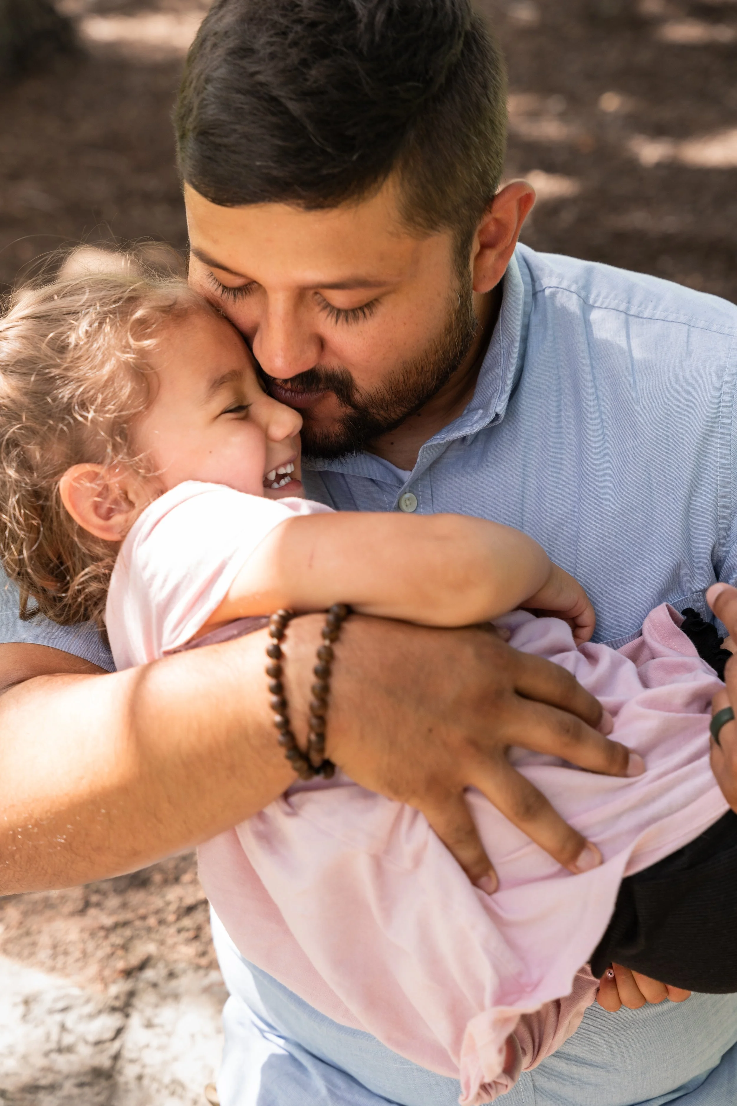 A man in a light blue shirt embraces a young girl with curly blonde hair, both smiling with their eyes closed, outdoors with dirt and trees in the background.