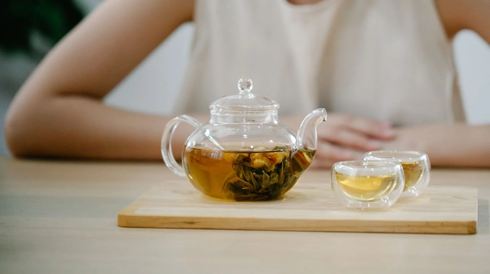 Glass teapot of golden green tea with opened leaves, beside two small glass cups on a wooden tray
