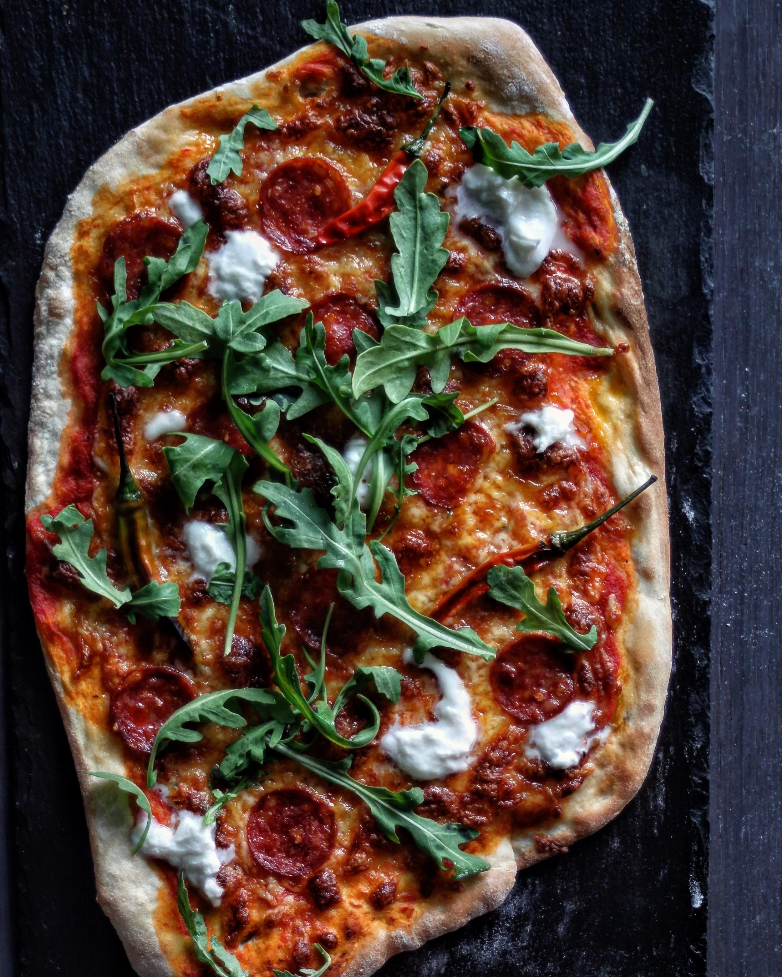 Ingredients 500g Strong White Flour 7g Yeast Sachet 1/2tbsp Caster Sugar 3tbsp Olive Oil 1/2tsp Salt 1 Tin Crushed Tomatoes 1 Clove Garlic Toppings (Pepperoni, Grated Mozzarella, Buffalo Mozzarella, Birdseye Chilis, Rocket) Method Begin by measuring out 325ml of tepid water into a jug, add the yeast, sugar, 2tbsp oil and stir to combine. Allow the mixture to sit for 5 minutes until bubbles begin to form on the surface. Sift the flour into a large mixing bowl and add the salt. When the yeast mixture is ready, slowly pour into the flour and use your hand to bring together into a dough. Tip out the dough onto a floured surface and kneed for 5-10 minutes until smooth and stretchy. Place into a clean bowl, cover with a damp tea towel and leave in a warm place for one hour or until doubled in size. Whilst the dough is rising, make the sauce by gently warming the tomatoes in a pan. Crush the garlic clove and add to the tomatoes with remaining oil and a good pinch of salt and pepper. Simmer for 5-10 minutes and set aside to cool. One hour before cooking the pizza, place a baking sheet or pizza stone into the oven and set the temperature to 220 degrees. Allow to get hot. When the dough is risen, knock back and divide into four portions. Roll each portion out on a lightly floured surface into a round (or any shape you like). Try and roll the dough thinly as this will achieve a crisper base. When the pizza stone or tray is ready and hot, place the pizza base directly on to it and top with 2-3 tablespoons of the sauce and spread over the base (leaving a small rim uncovered for the crust). Top the pizza with cheese and your preferred toppings, a drizzle of extra virgin olive oil and place into the oven for 10-12 minutes or until golden, crisp and bubbling. Serve immediately topped with fresh rocket.