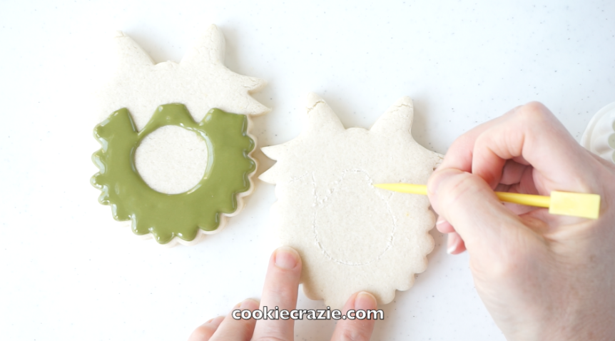  Once the cookie is baked and cooled, use a  boo boo stick  or toothpick to etch lightly on the cookie surface, making a circle in the middle and an outline of the bow portion. [This will give you a reference of where to pipe the glaze.] 