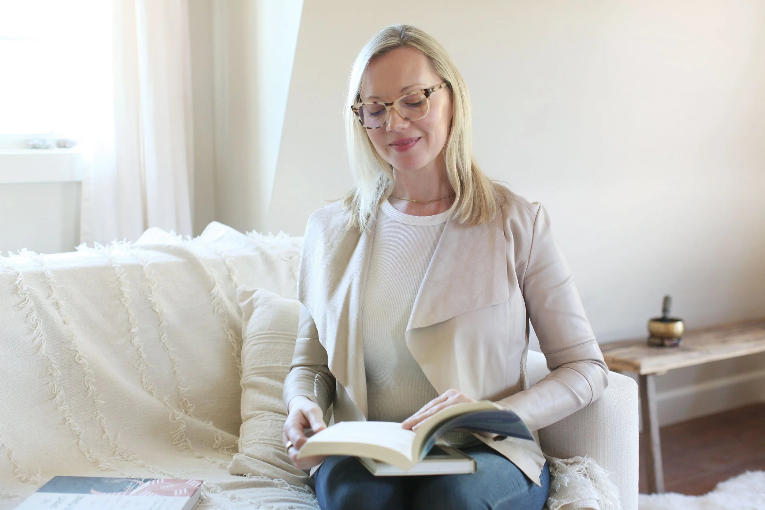 Woman sitting on a beige sofa in a bright room, reading a magazine with a book on her lap, wearing glasses and a light-colored blazer.