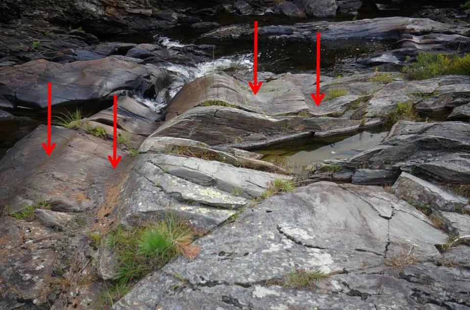 Figure 1: Several parallel glacial grooves are identified by the red arrows. The glacial grooves were created by the slow movement of the massive glacier that once covered almost all of Canada. As glacier slowly moved, or flowed, across the rock land, the glacier scoured troughs, of furrows, into the bedrock. The rock and grooves are exposed in the Aux Sables River, Chutes Provincial Park, Ontario, Canada. Photo by Andy Fyon, Sept 11/18.