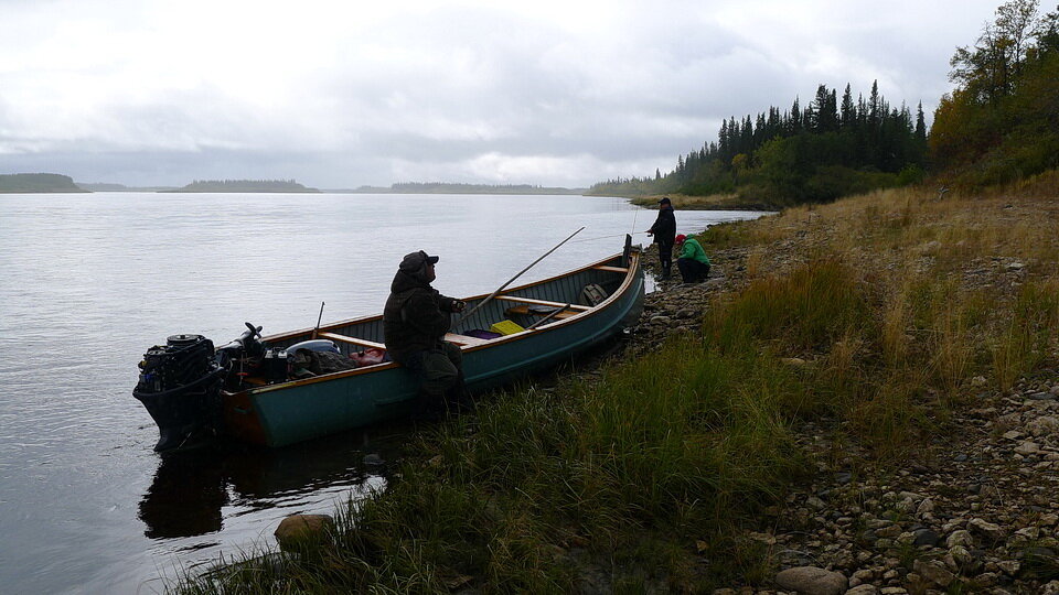 图1:维克多·瓦巴诺(在货轮独木舟上喝着必喝的茶)、苏珊娜(本名哈莱特;绿夹克)和Ivan Iserhoff在Winisk河岸边钓鱼，Peawanuck北部，安大略省，9月21日至12日。raybet11Andy Fyon摄于9月21日至21日。
