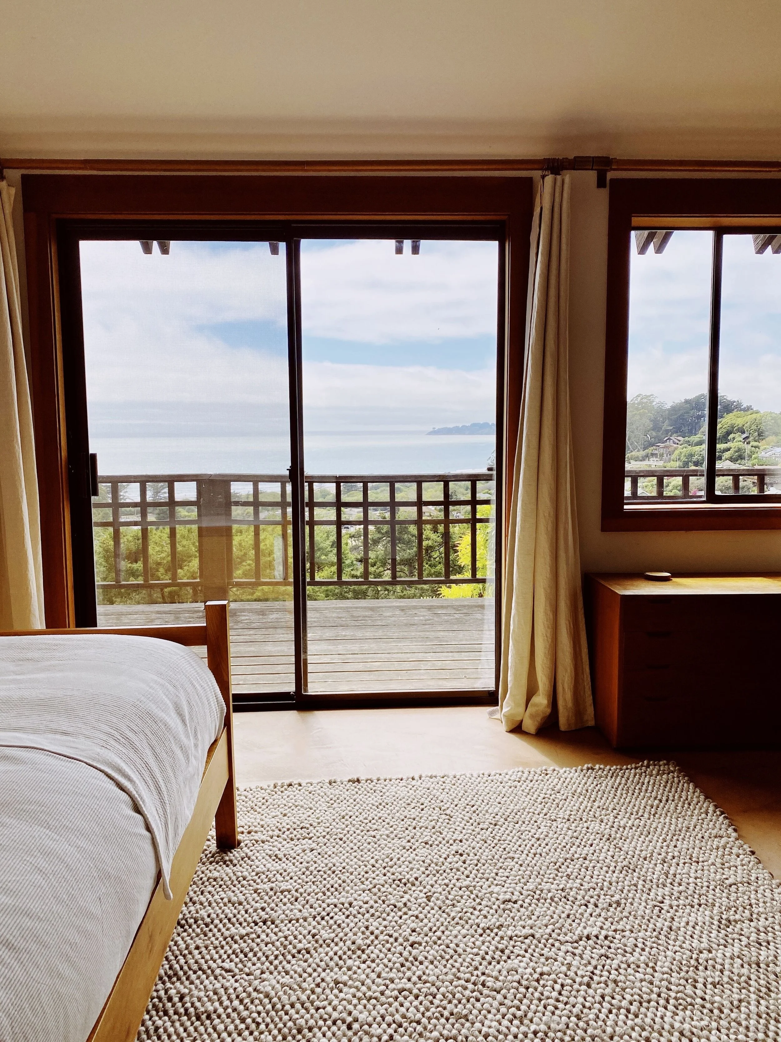 Room with sliding glass door leading to a balcony, view of trees, ocean, and sky outside. 