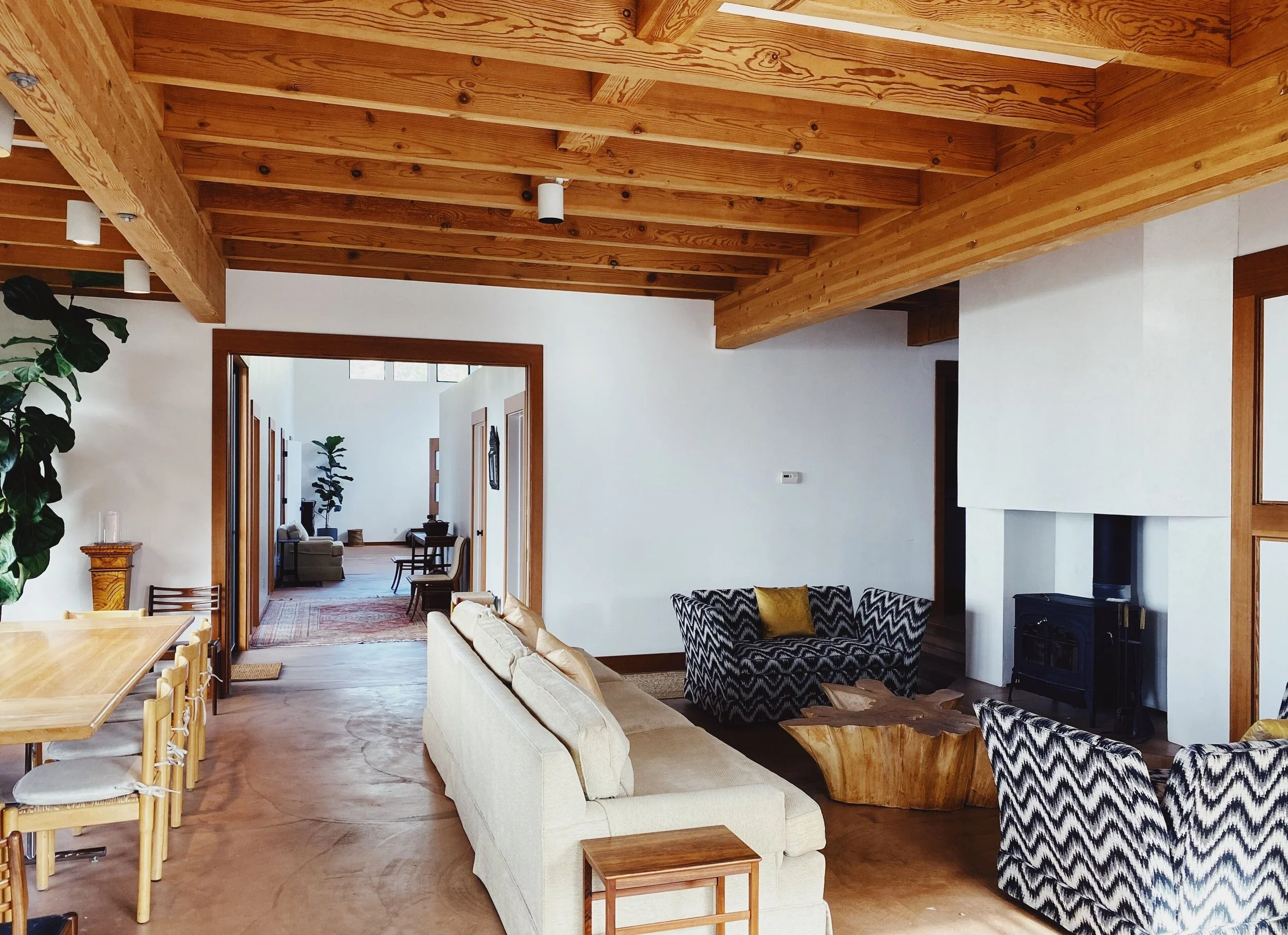 Interior living room with wooden ceiling beams, white walls, beige sofa, patterned armchairs, coffee table made from natural wood, fireplace, and additional seating in the background.