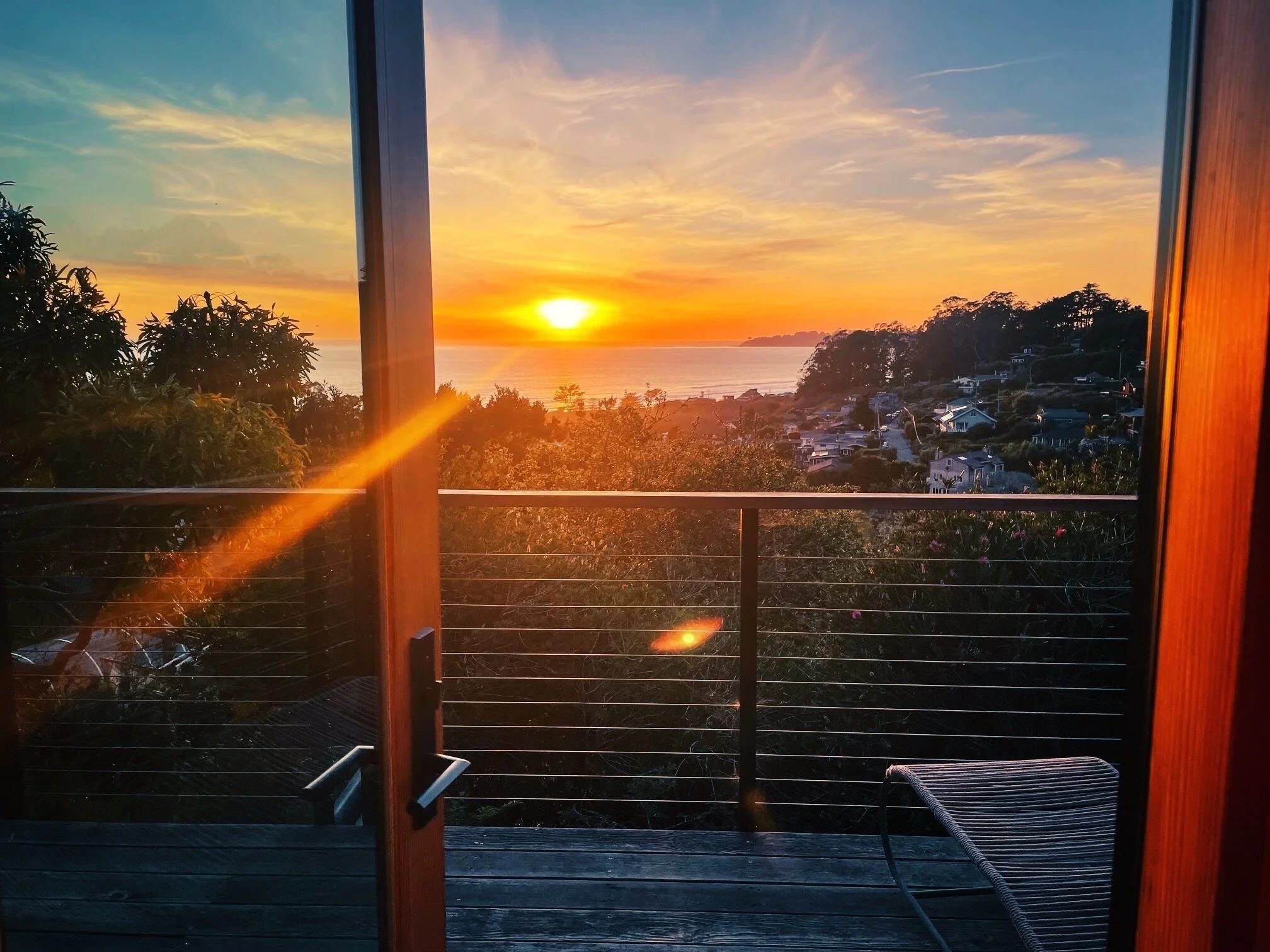 Sunset view through open doorway with a balcony railing, trees, and houses in the distance over water.