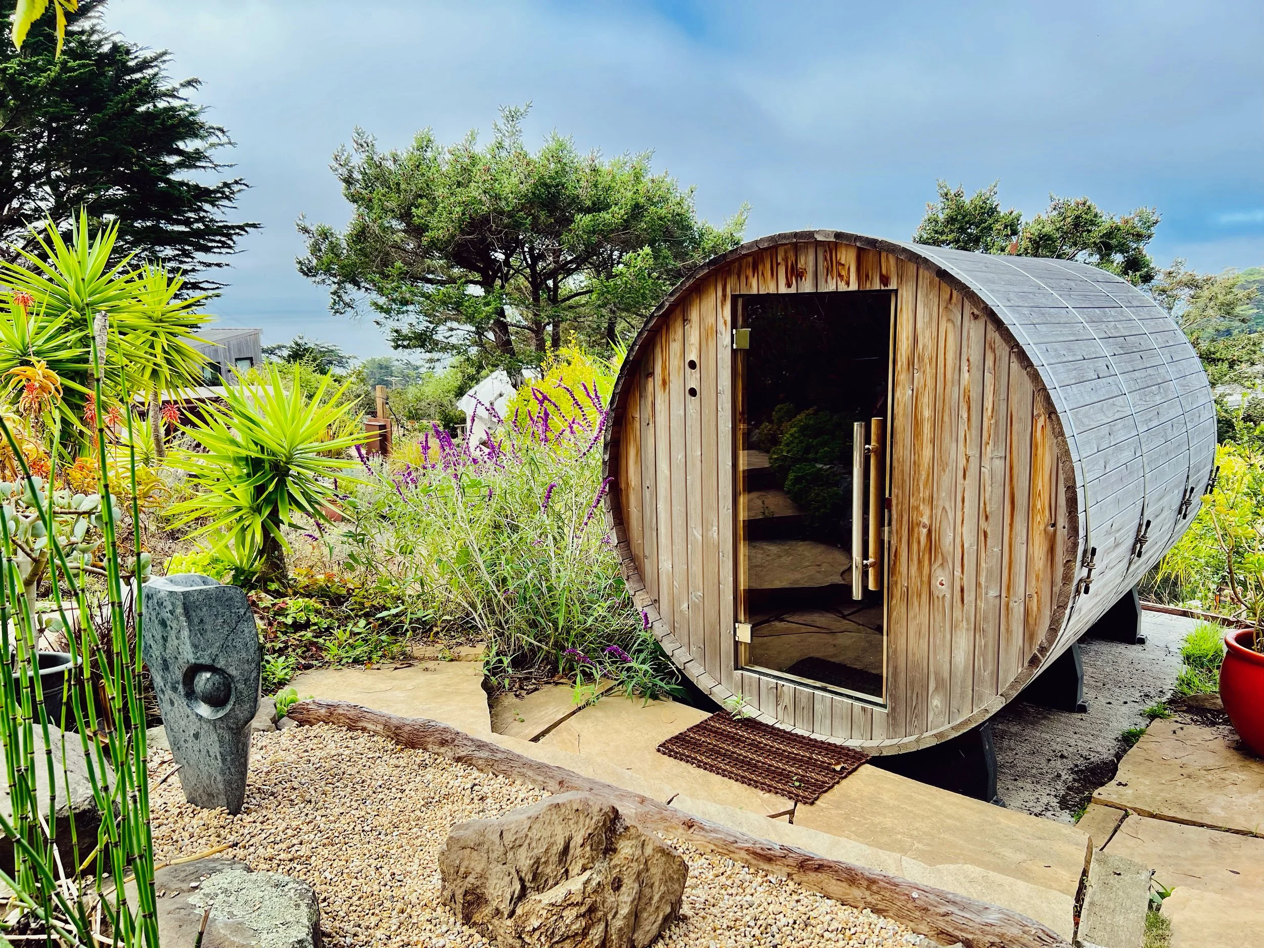 A wooden barrel-shaped sauna  in a lush garden. A space to reset the nervous system. 