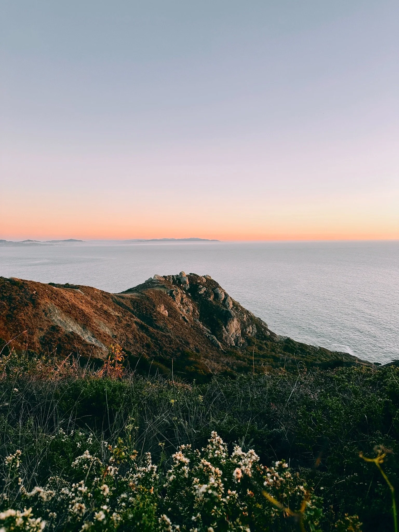 Coastal landscape with rocky hills, green vegetation, and a calm ocean under a pastel sunset sky.