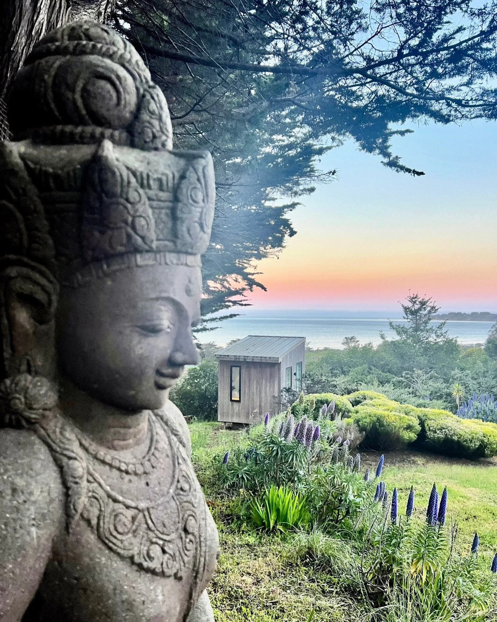 A stone sculpture of a smiling deity with detailed carvings, surrounded by lush green plants and flowers, overlooking the ocean at sunset.