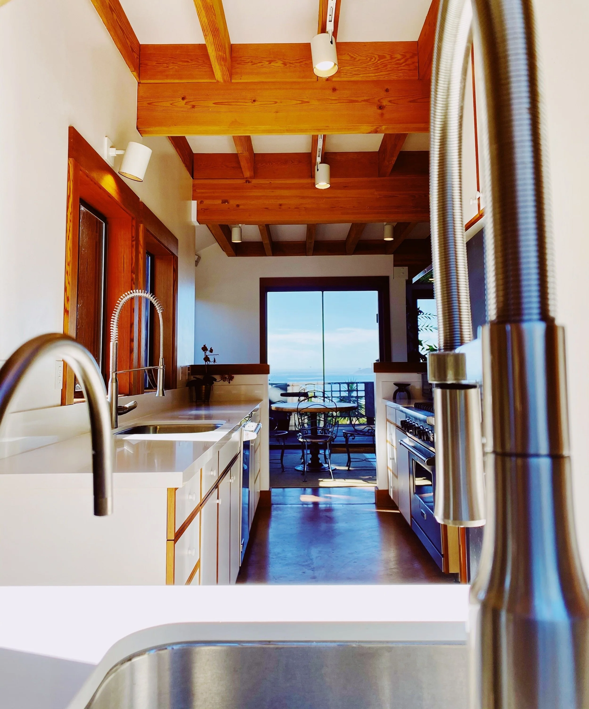 A view from the kitchen sink looking towards a bright room with wooden ceiling beams, large glass doors leading to a balcony with outdoor seating, and a scenic ocean view in the distance.