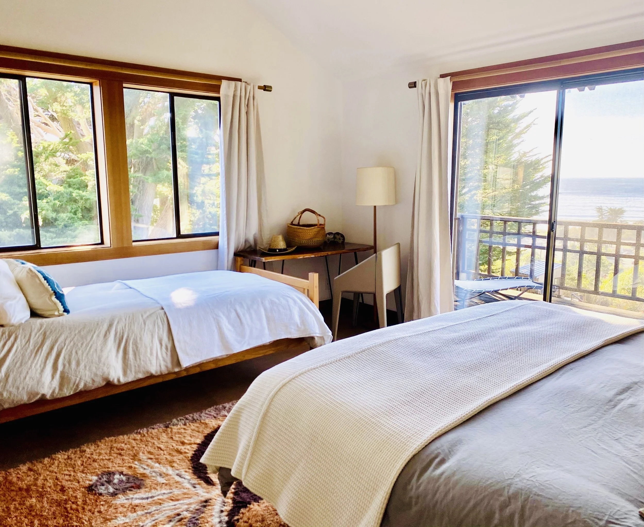 A coastal bedroom with two beds, large windows, a sliding glass door leading to a balcony, and ocean view.